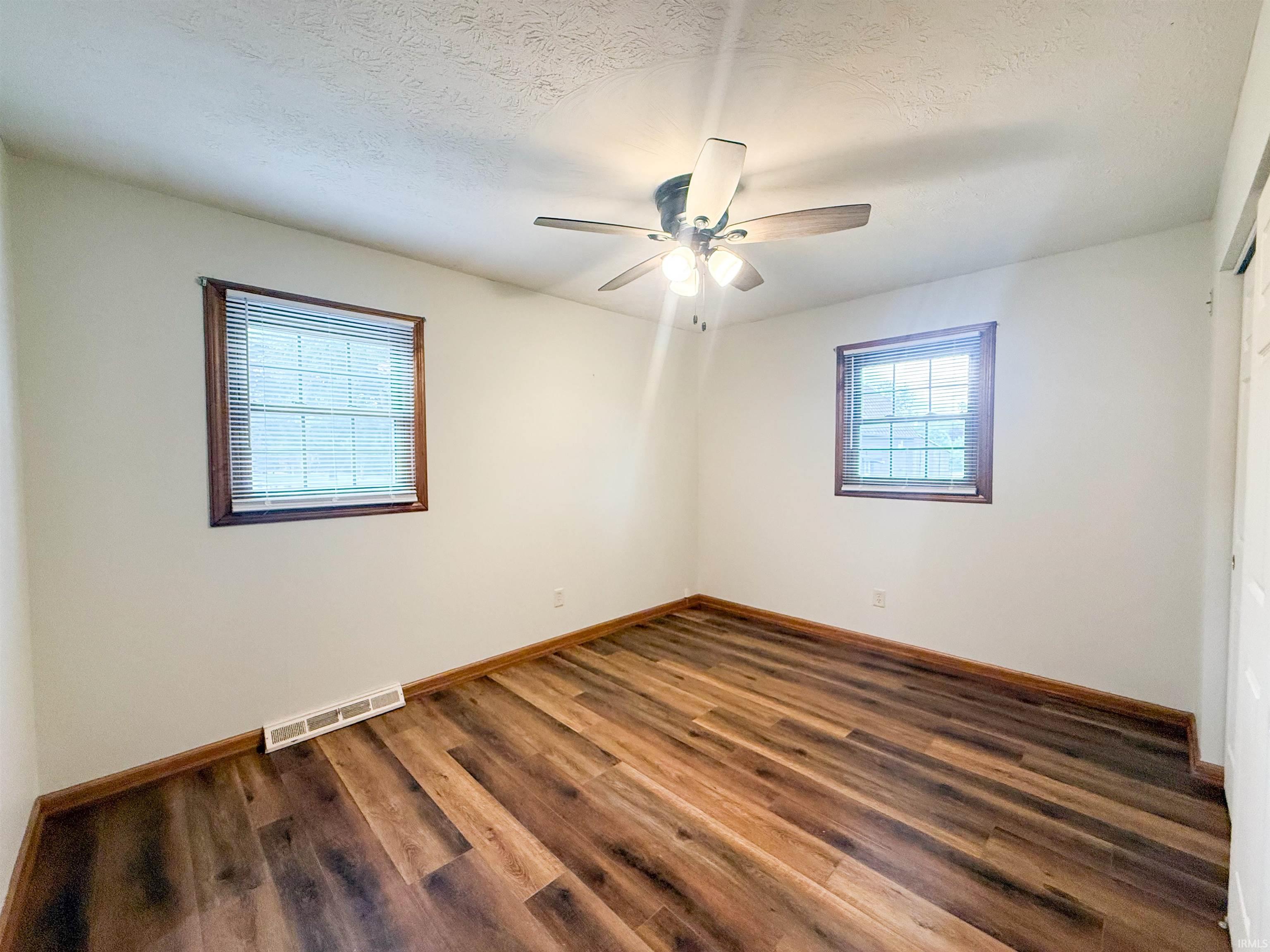 Bright interior space with wood-finish flooring, two windows with blinds, a ceiling fan with integrated lighting, and white painted walls