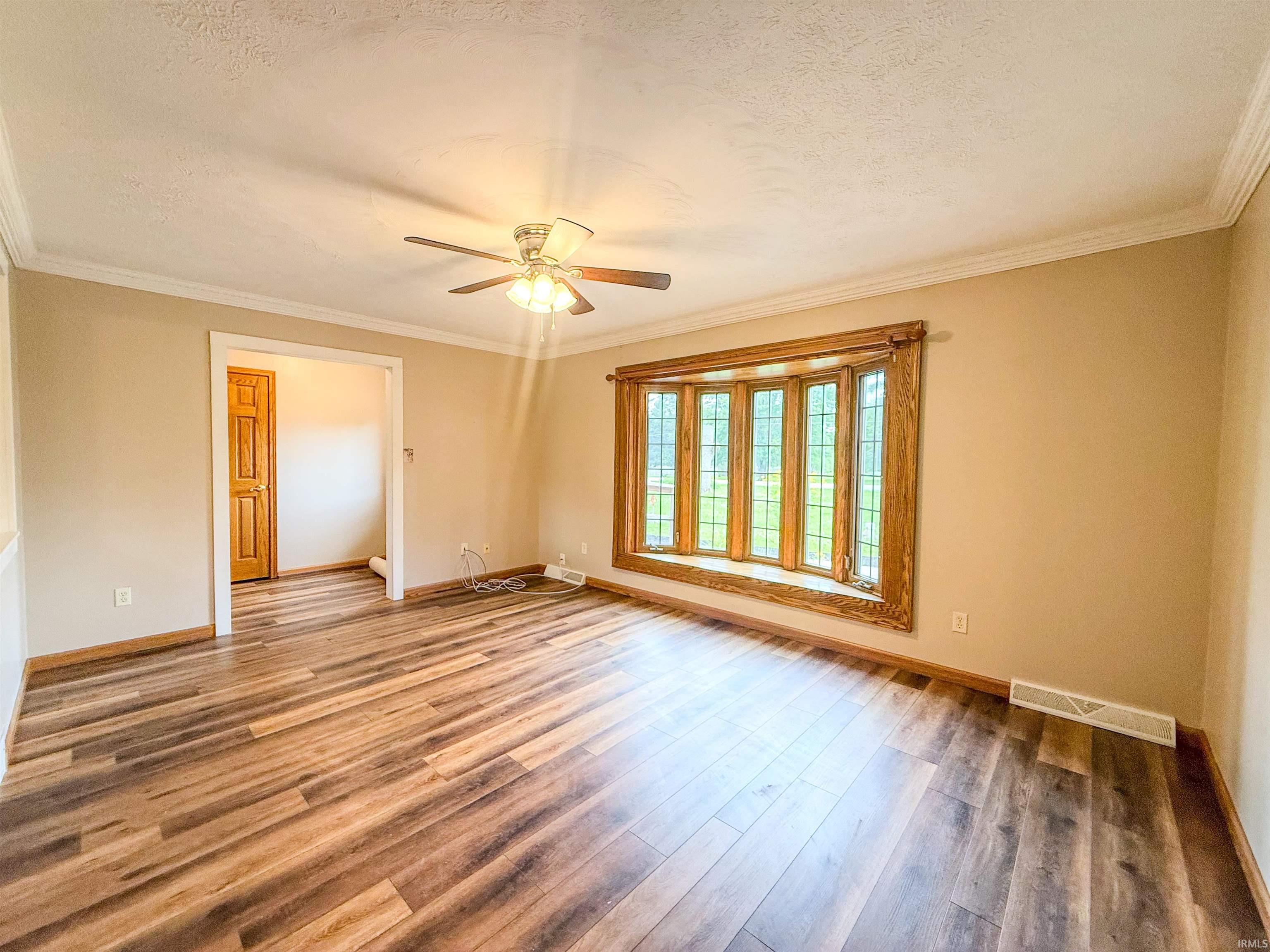 Spacious room featuring wood-finish flooring, a large bay window with wood trim, a ceiling fan with light fixture, white crown molding, and a warm neutral paint palette
