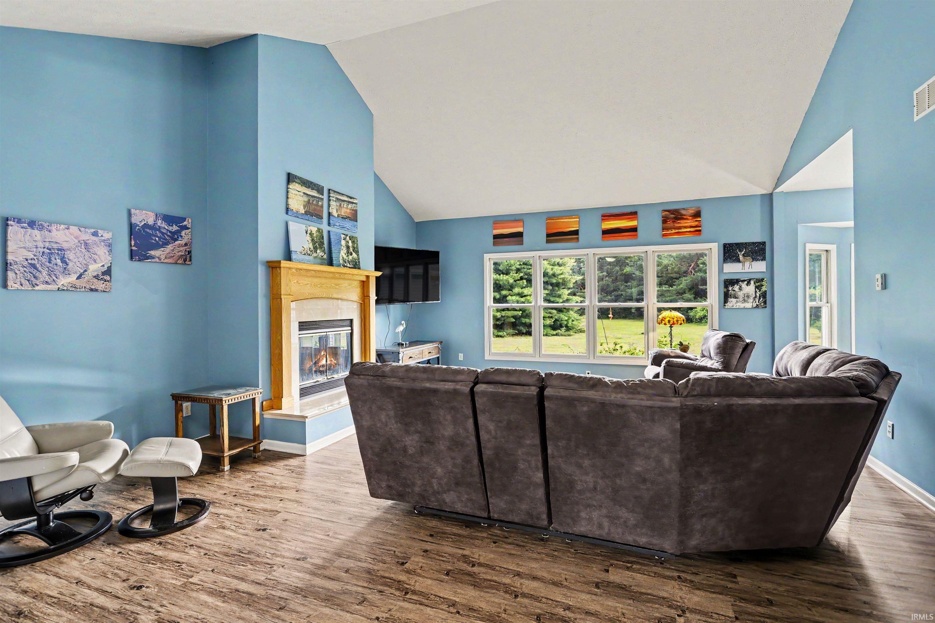 Spacious living area featuring a vaulted ceiling, wood-finish flooring, a fireplace with a light-toned wood mantel and stone surround, and multiple windows providing outdoor views
