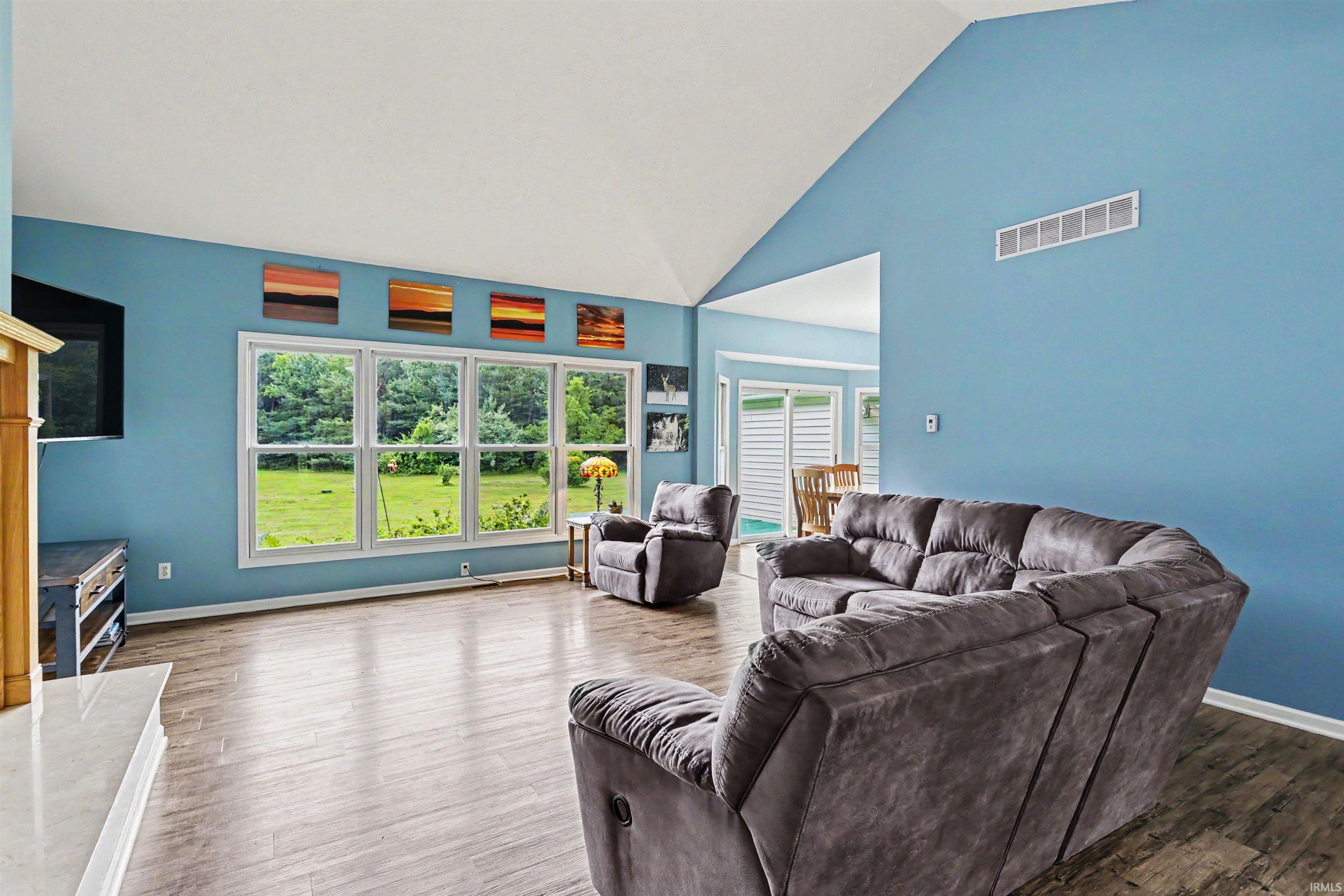 Spacious living room featuring a vaulted ceiling, wood-finish flooring, a white mantel, and a multi-panel window overlooking a green landscape