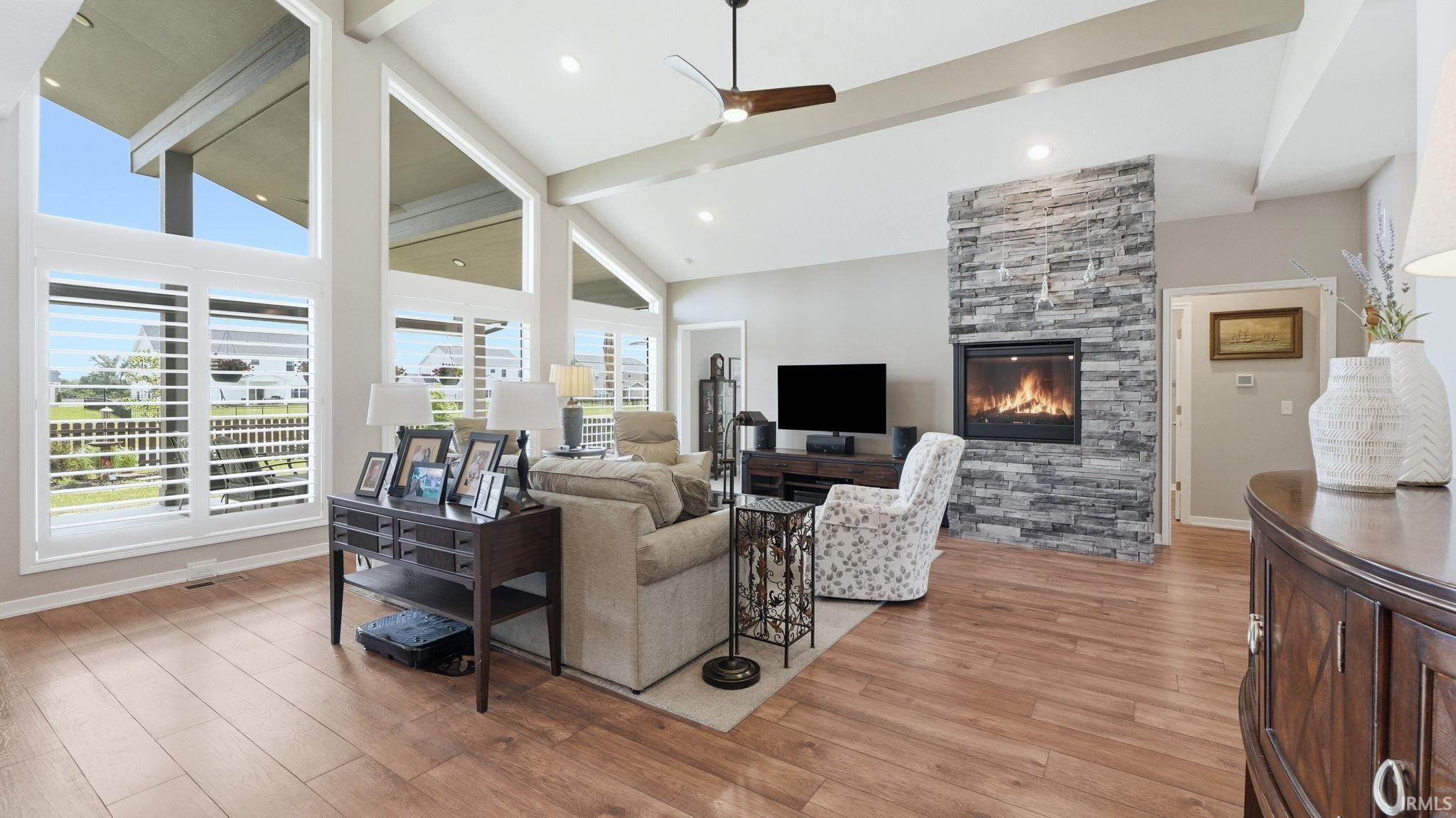 Spacious living area featuring a stone-clad fireplace, vaulted ceilings with exposed beams, wood-finish flooring, a ceiling fan, and large windows with integrated plantation shutters
