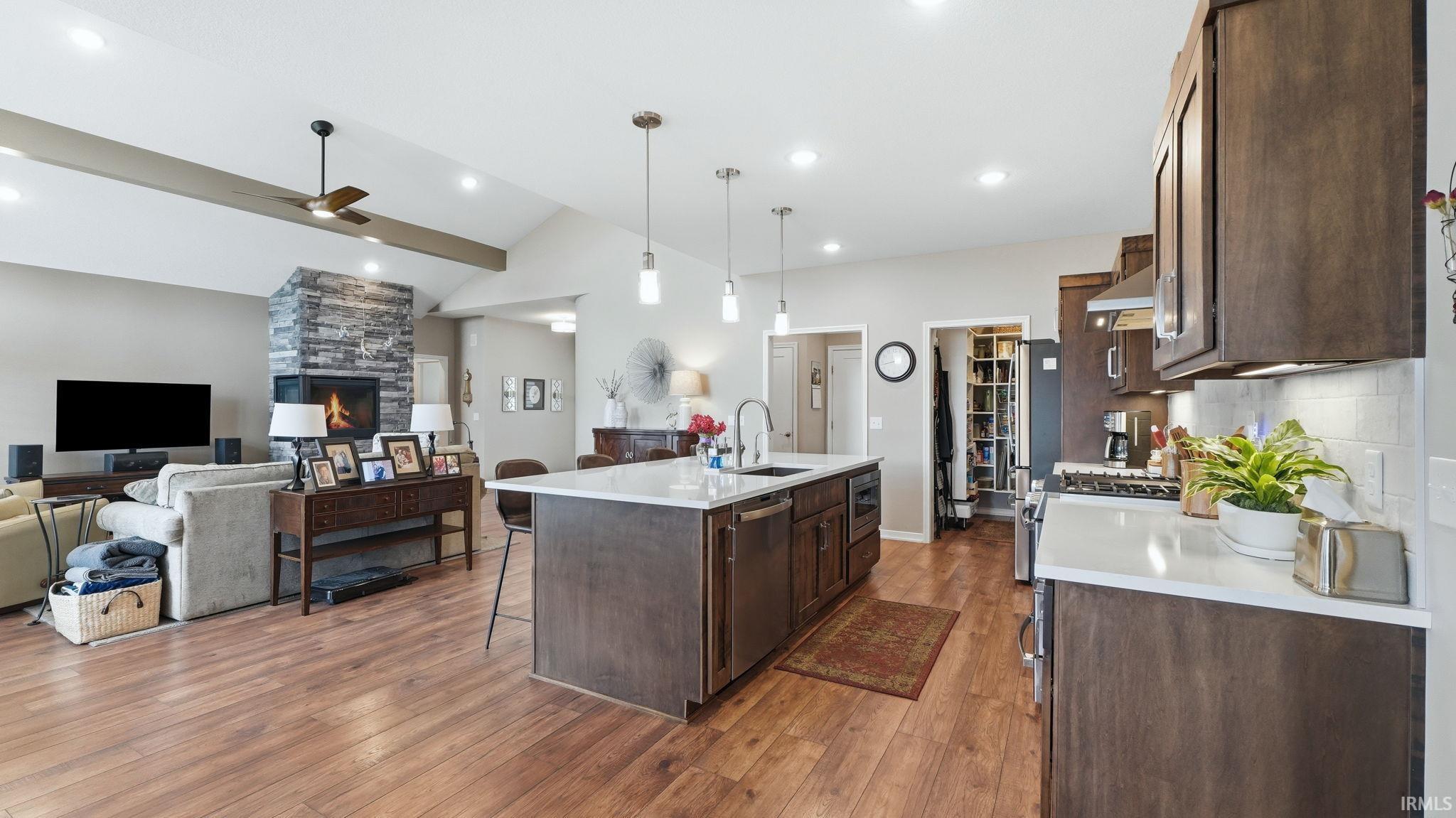Kitchen and living area featuring wood-finish flooring throughout, a central island with a white countertop and pendant lighting, built-in dark wood cabinetry, a stone-front fireplace, and vaulted ceilings