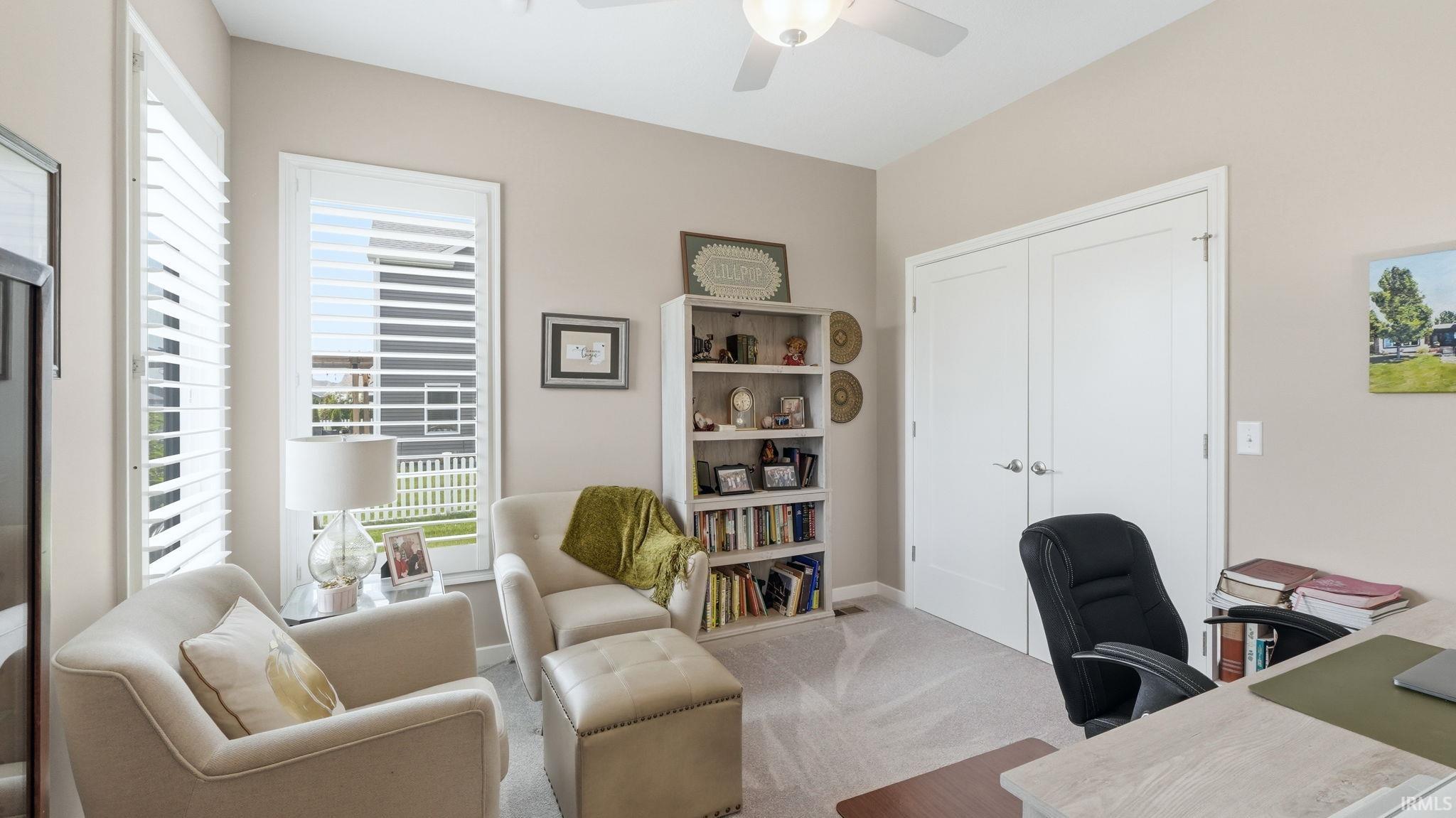 Neutral-toned interior room featuring twin windows with white plantation shutters, a ceiling fan with integrated lighting, and a built-in white bookshelf