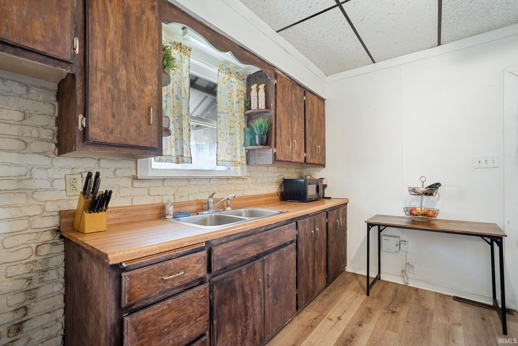 Kitchen featuring wood-finish cabinetry, a double basin stainless steel sink, wood-finish countertops, a white brick-style backsplash, and wood-finish flooring