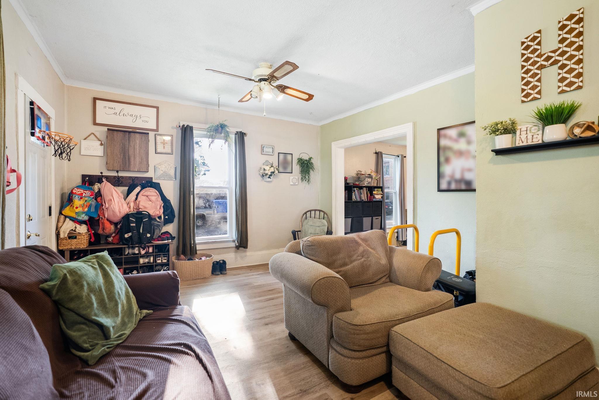 Living area featuring wood-finish flooring, a ceiling fan with light fixture, natural light from multiple windows, and crown molding