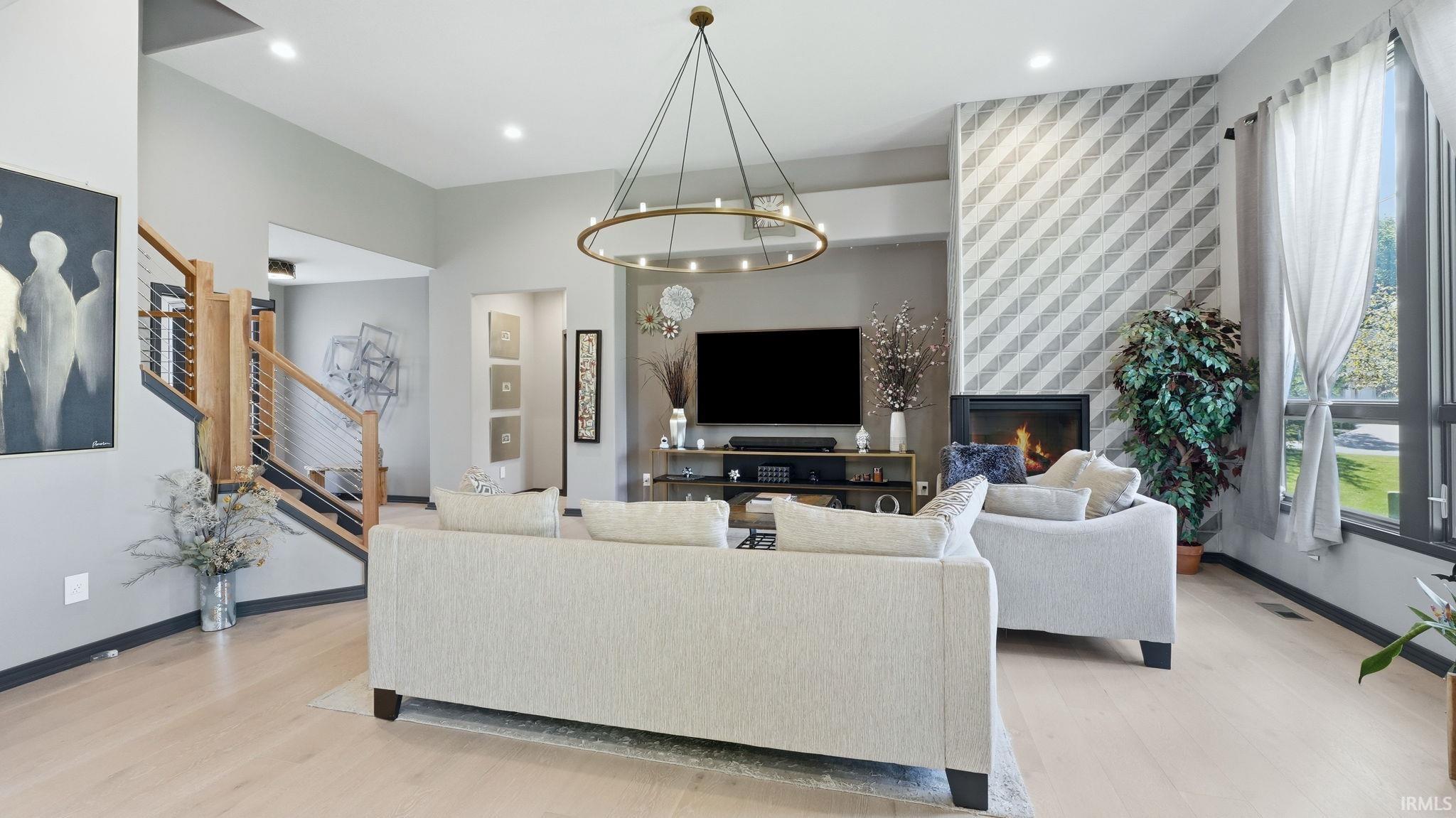 Spacious living area featuring light wood-finish flooring, a contemporary chandelier, a fireplace with a decorative accent wall, recessed lighting, and a modern staircase with wood and metal railing