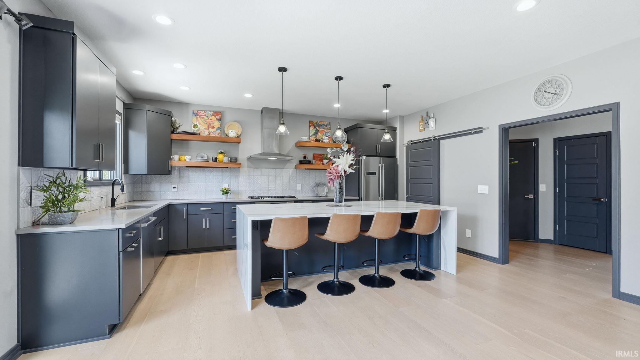 Modern kitchen featuring dark gray cabinetry, light wood-finish flooring, a large central island with seating, a stainless steel range hood, and open wooden shelving
