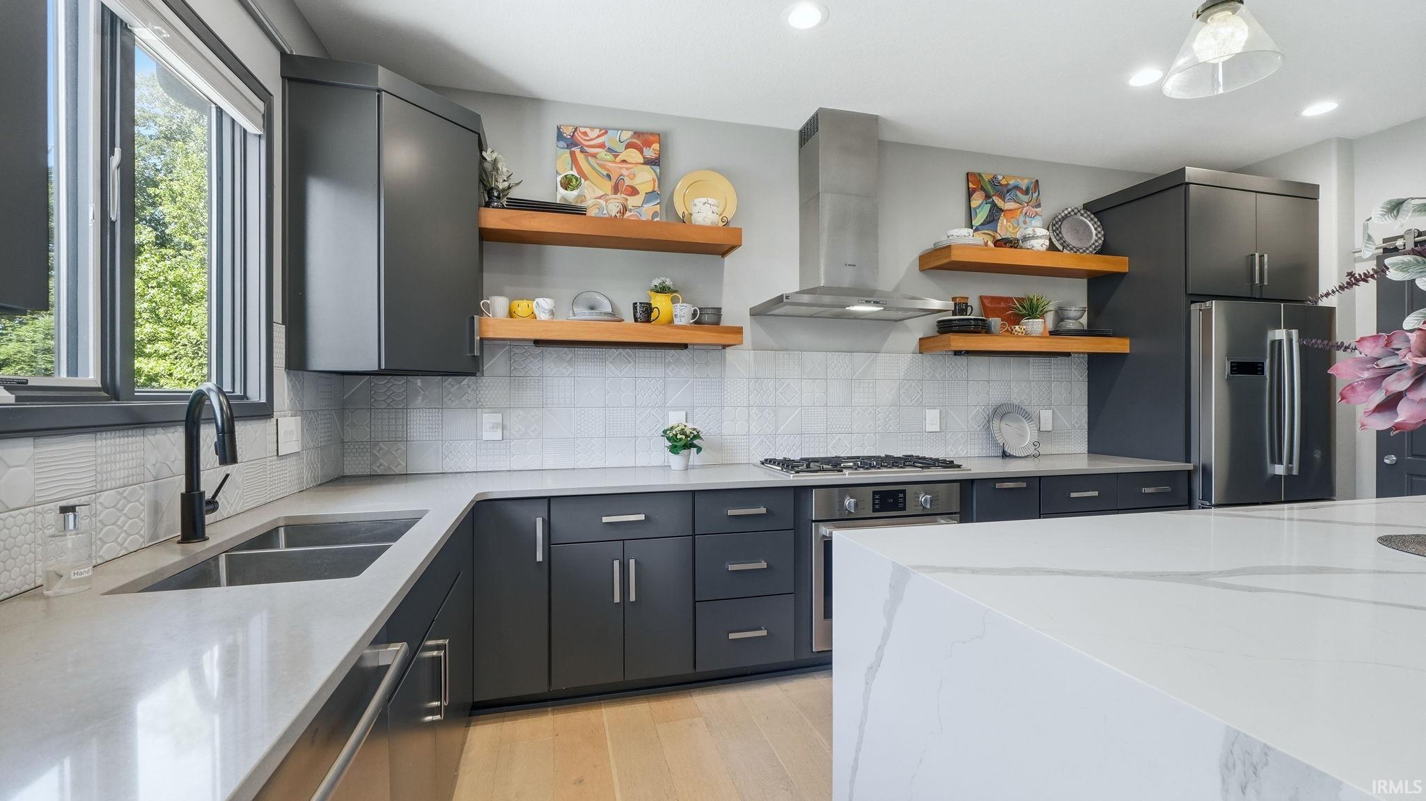 Contemporary kitchen featuring dark cabinetry, light-toned countertops, a stainless steel range hood, floating wood shelves, and light wood-finish flooring