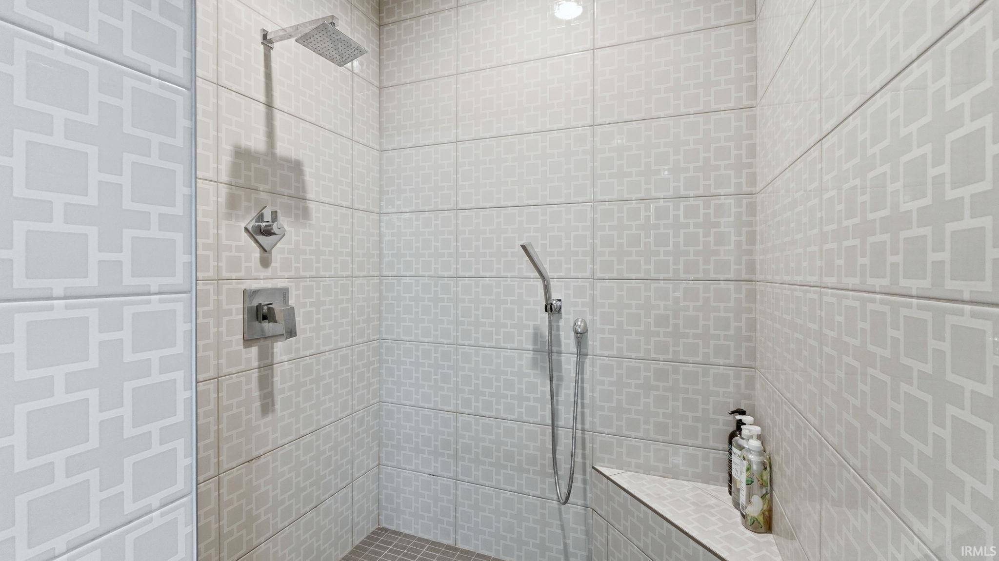 Walk-in shower featuring patterned tile walls, a built-in shower bench, a rain showerhead, a handheld shower fixture, and polished chrome hardware
