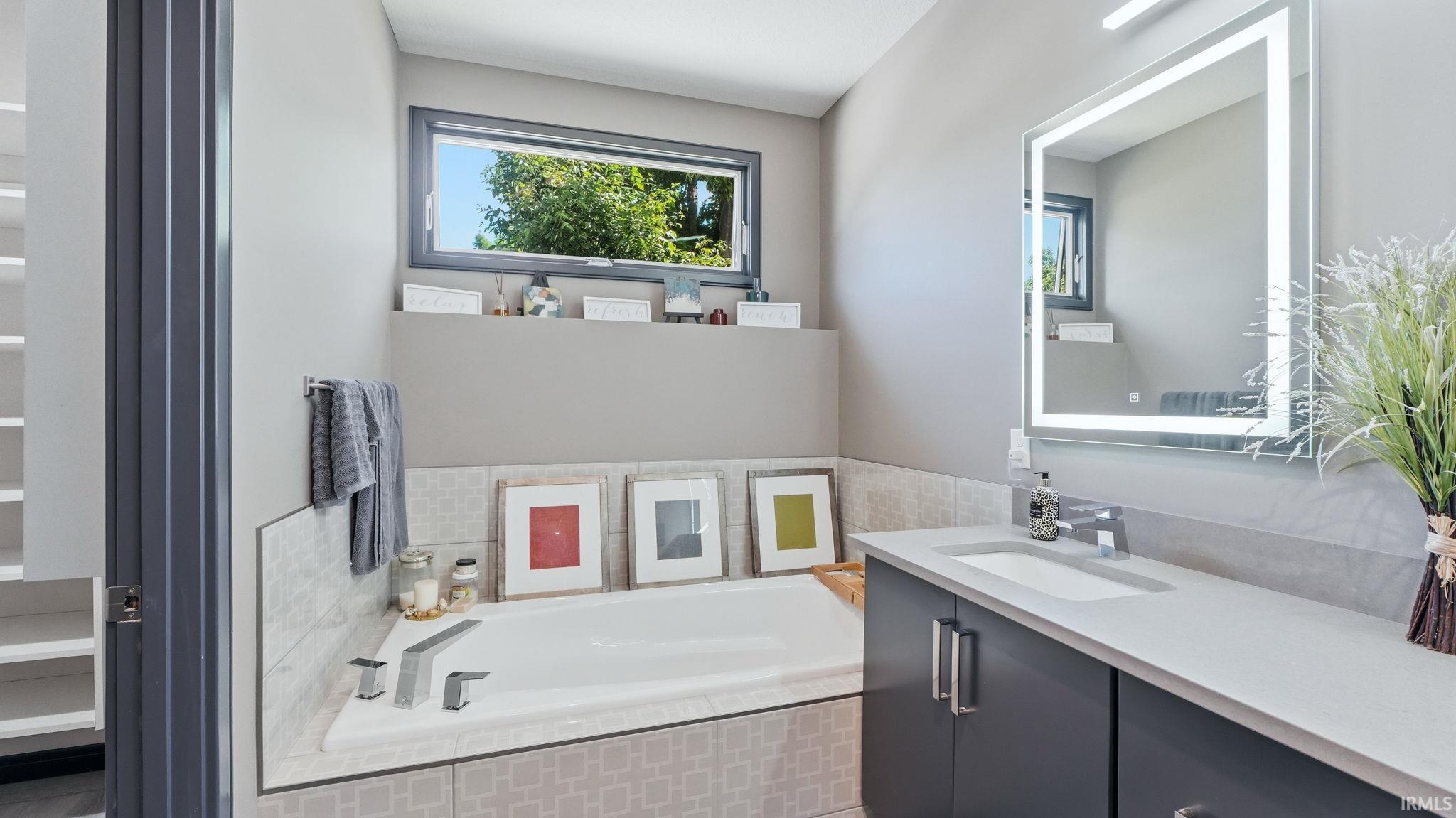 Contemporary bathroom featuring a built-in soaking tub, an integrated vanity sink, a large wall-mounted illuminated mirror, dark cabinetry with chrome hardware, and a high horizontal window