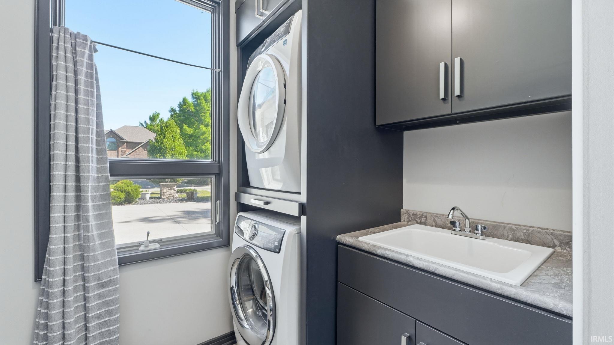 Laundry area featuring a stacked washer and dryer, dark cabinetry with brushed nickel hardware, an integrated utility sink, and a framed window