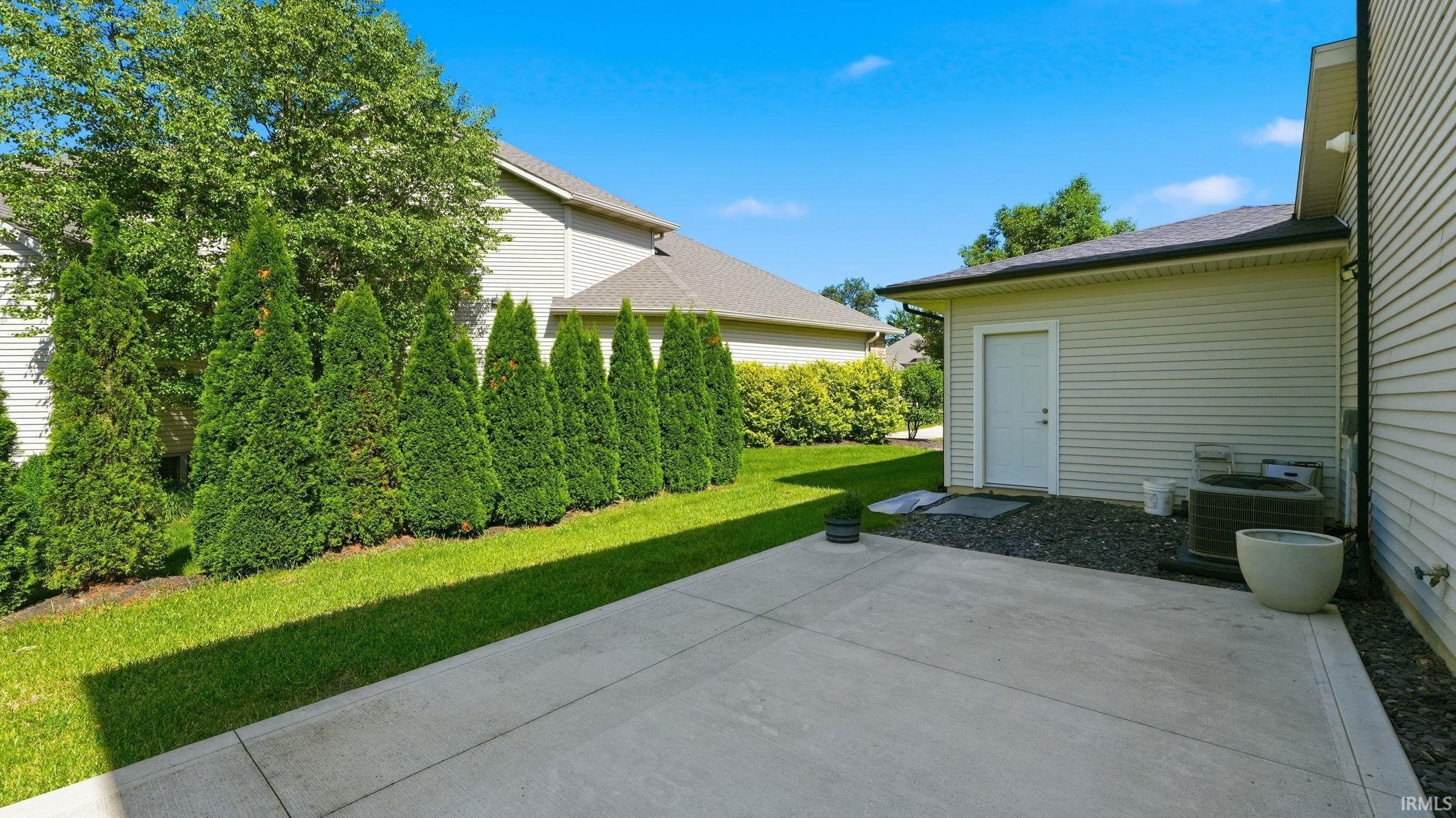 Concrete patio area featuring an exterior door, gravel landscaping, and a row of mature evergreen trees