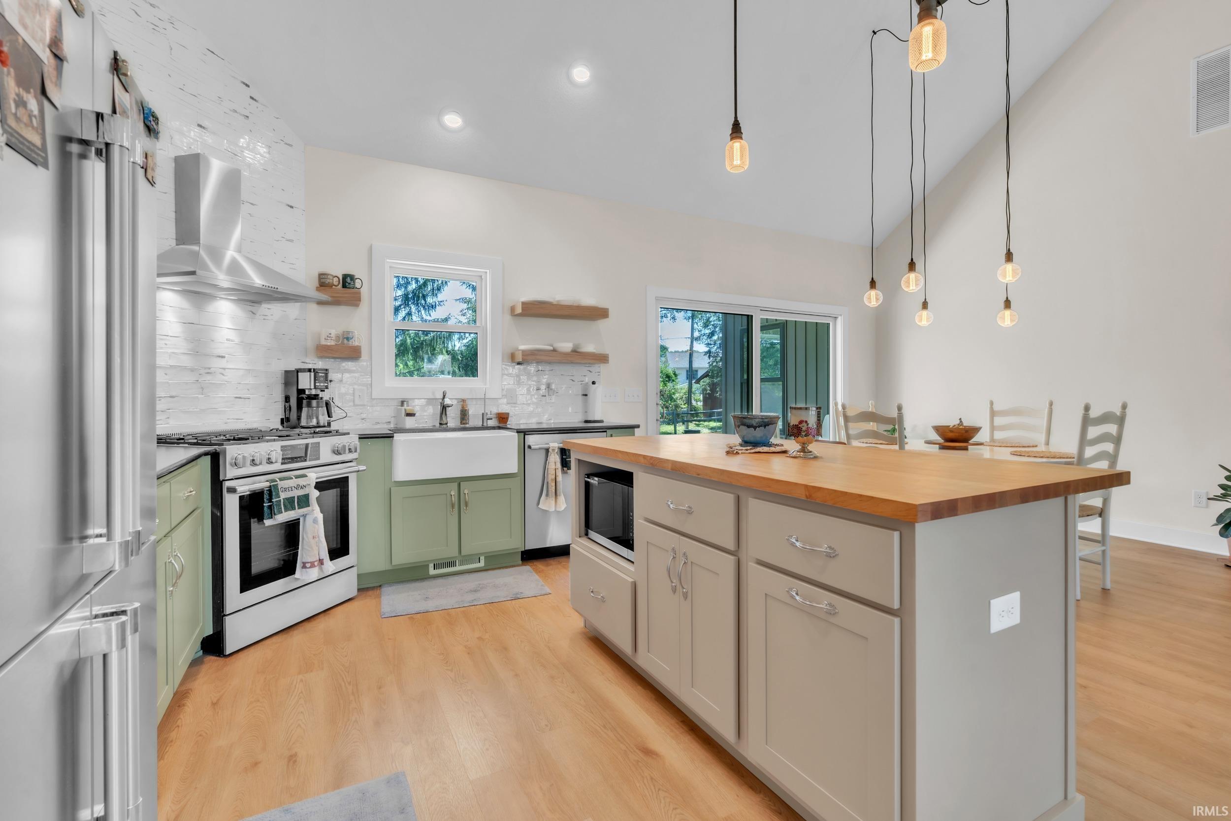 Kitchen featuring a large central island with a wood-finish countertop, stainless steel appliances, a farmhouse sink, light green base cabinetry, and wood-finish flooring