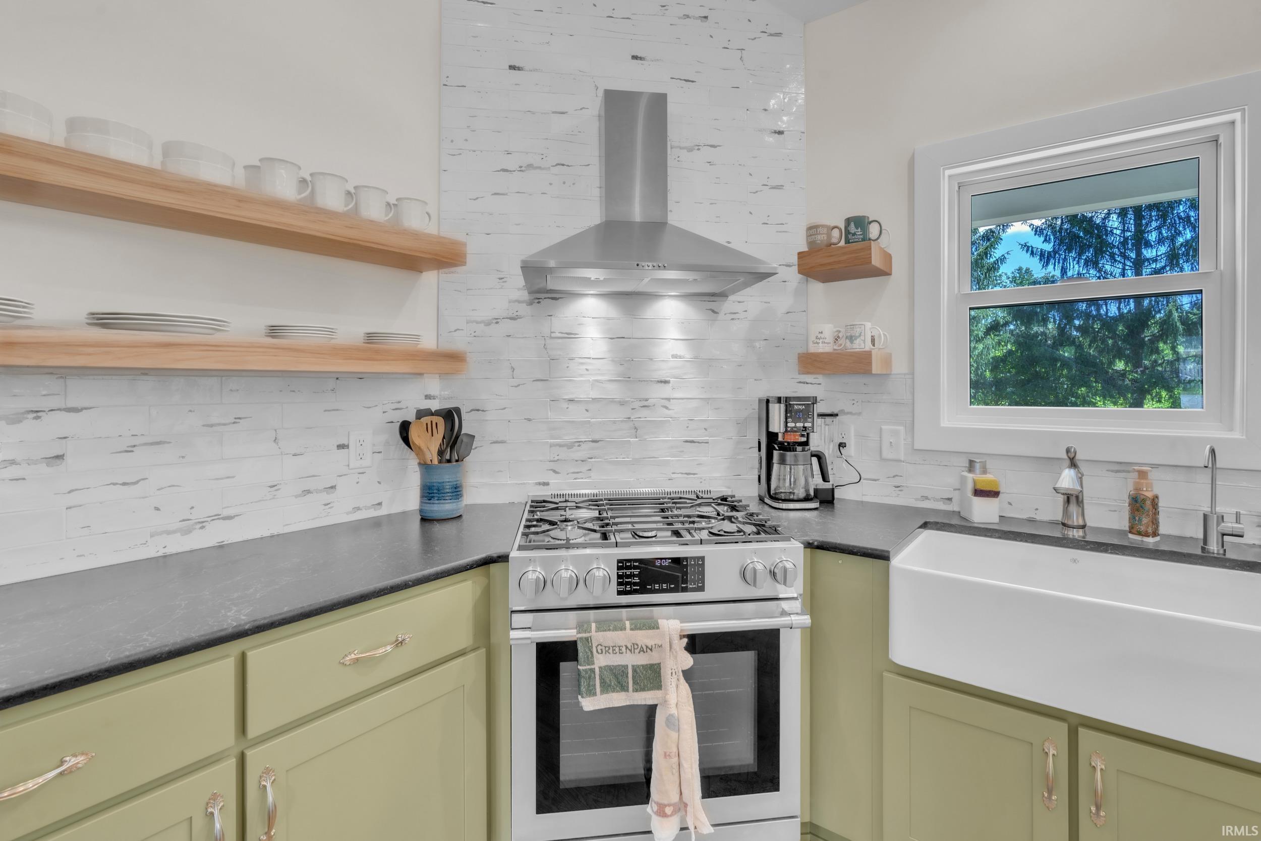 Kitchen featuring a stainless steel range with a coordinating range hood, a white farmhouse sink with a gooseneck faucet, light green cabinetry, dark countertops, and a white tile backsplash