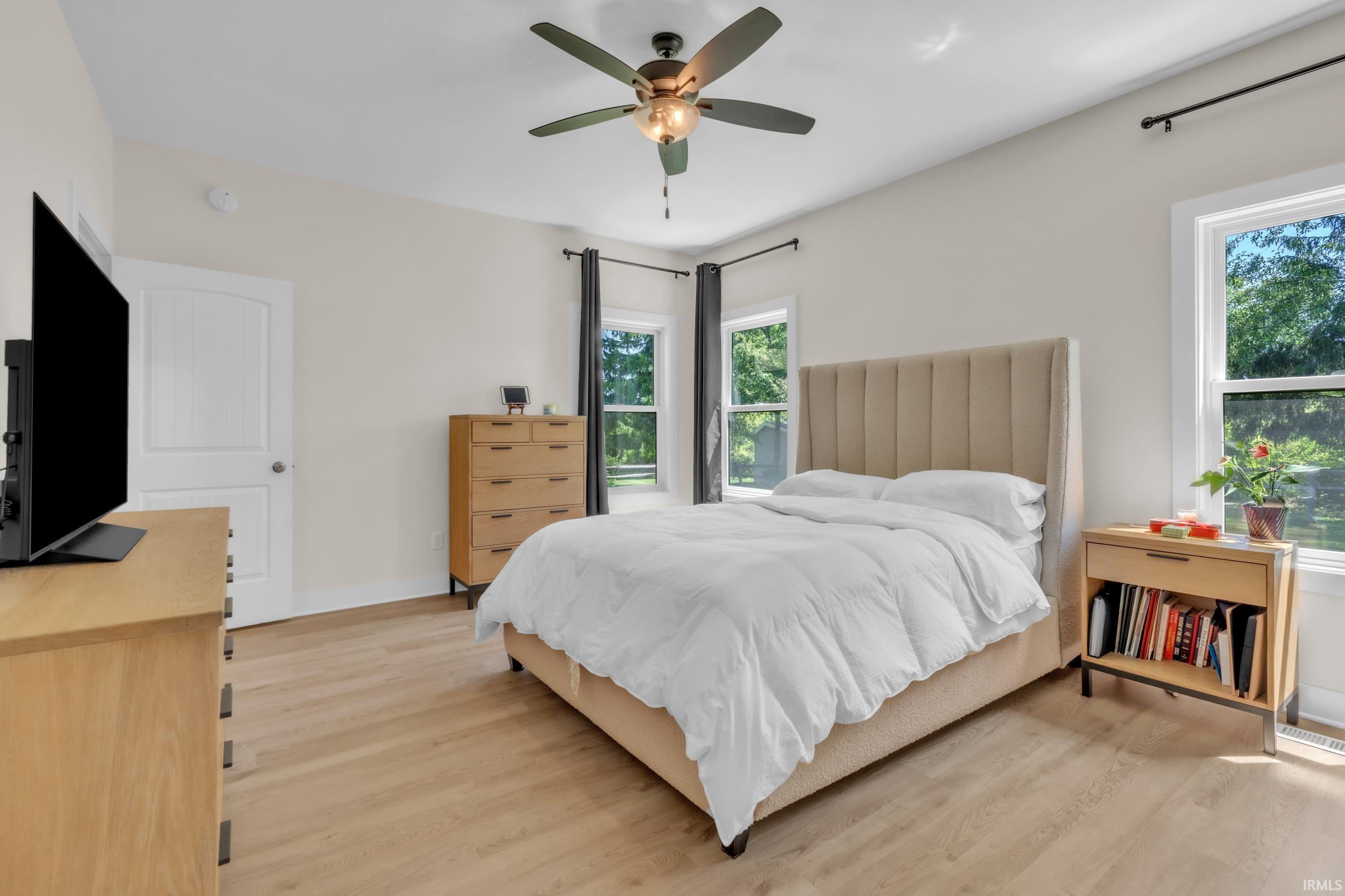 Bedroom featuring wood-finish flooring, a ceiling fan with integrated lighting, two windows with dark curtain rods, and a white paneled door