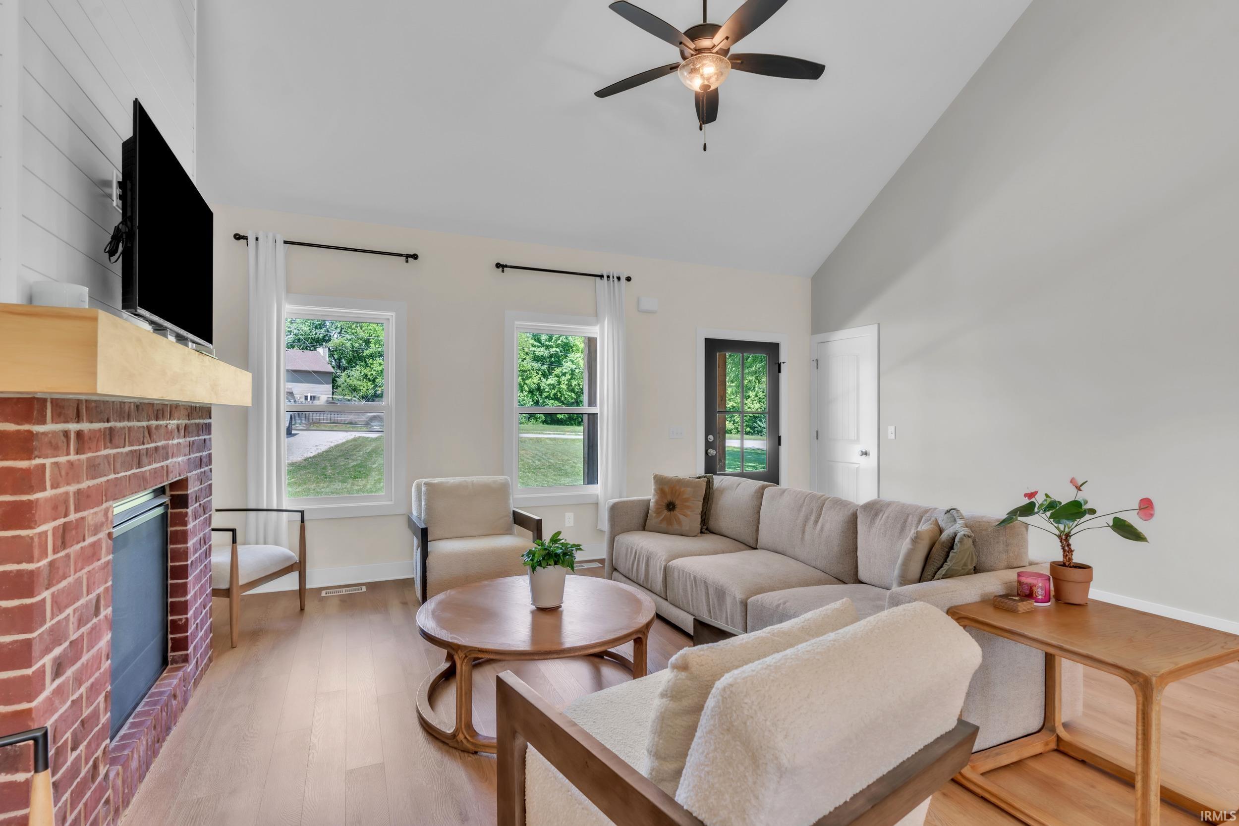 Vaulted ceiling with a ceiling fan, brick fireplace with a wood mantel, wood-finish flooring, and multiple windows