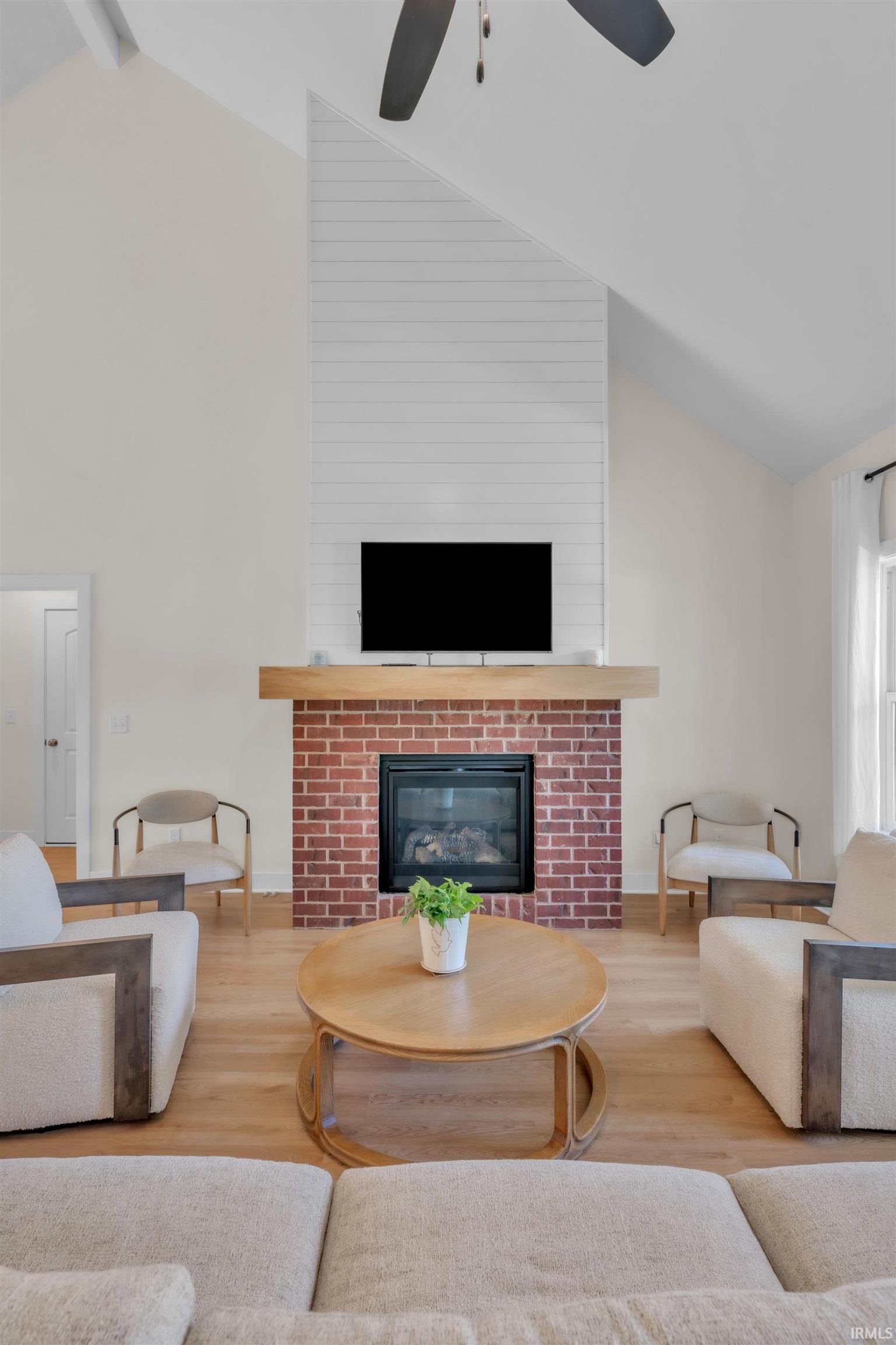 Vaulted living area featuring a brick fireplace with a wood mantle, shiplap accent wall, wood-finish flooring, and a ceiling fan