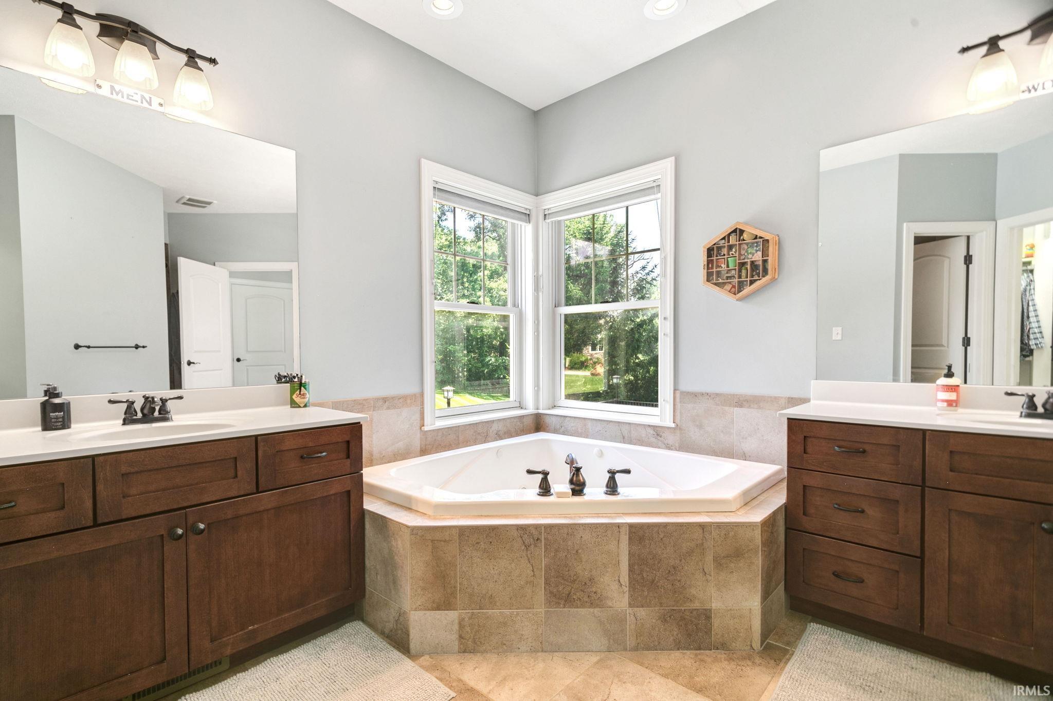 Corner soaking tub with tiled surround, dual wood-finish vanities with solid surface countertops, and windows providing ample natural light