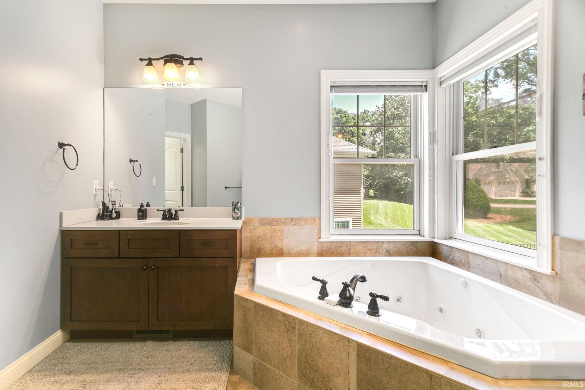 Soaking tub with jetted features and an expansive window, complemented by a wood-finish vanity with dual sinks, a large mirror, and overhead lighting