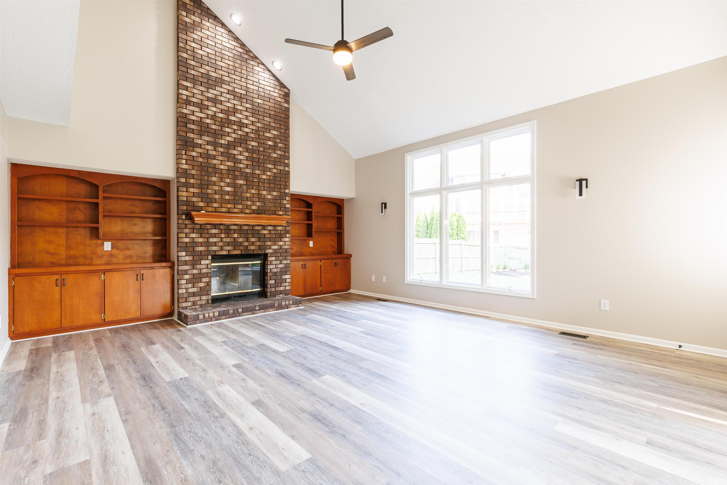 Spacious living area featuring a vaulted ceiling, prominent brick fireplace with a wood mantle, built-in wooden cabinetry, multi-pane windows, and wood-finish flooring