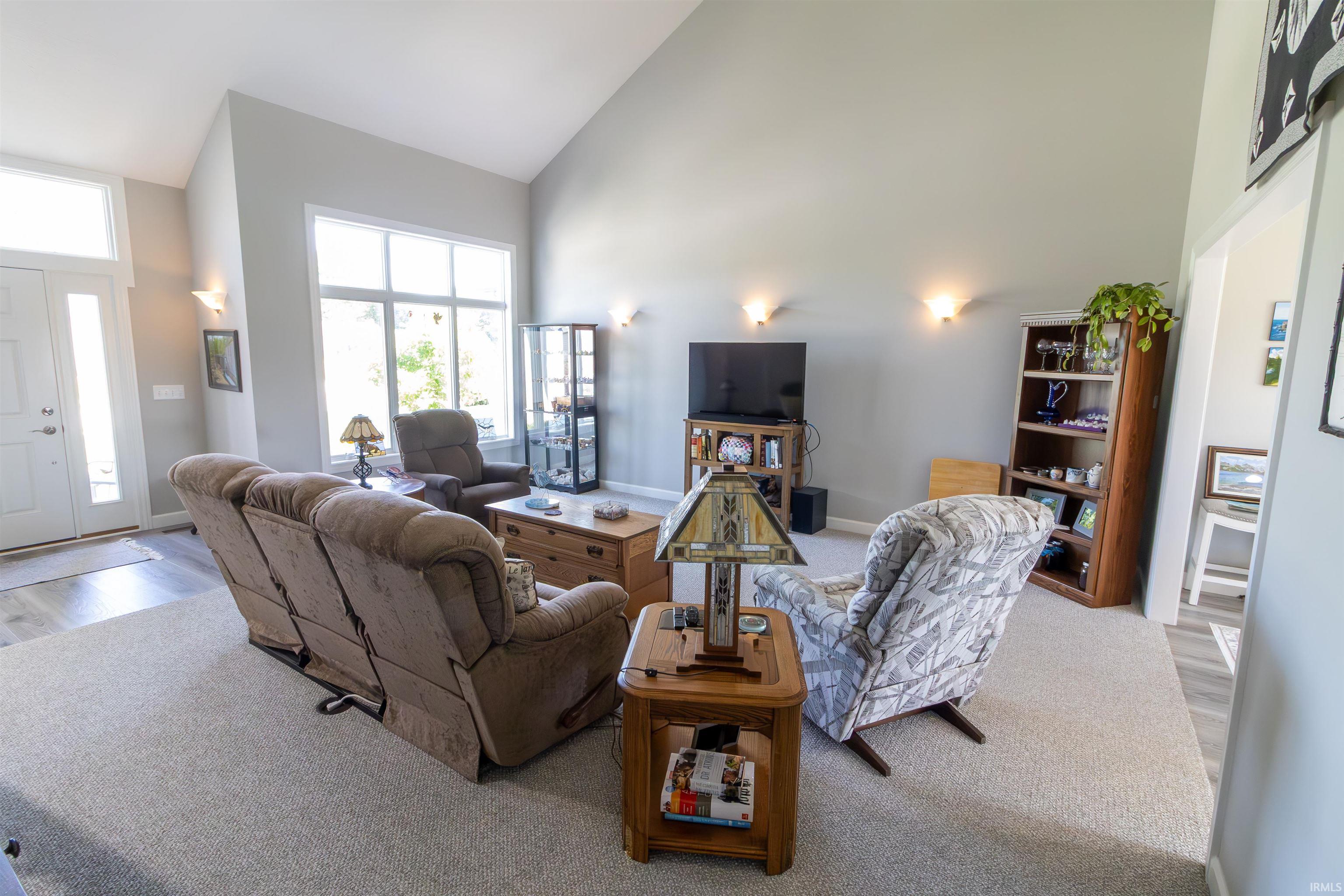 Vaulted ceiling living area featuring a large picture window, light gray wall tones, wood-finish flooring at the entry, and wall sconce lighting