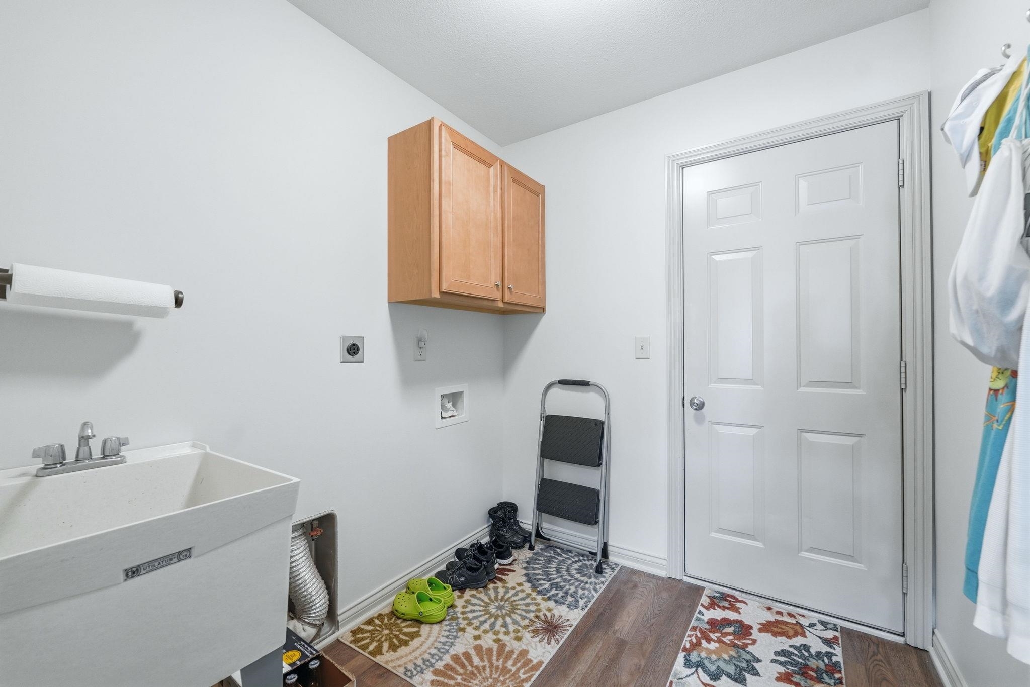 Dedicated laundry space featuring wood-finish flooring, a utility sink with dual-handle faucet, natural wood cabinetry, and a white paneled door