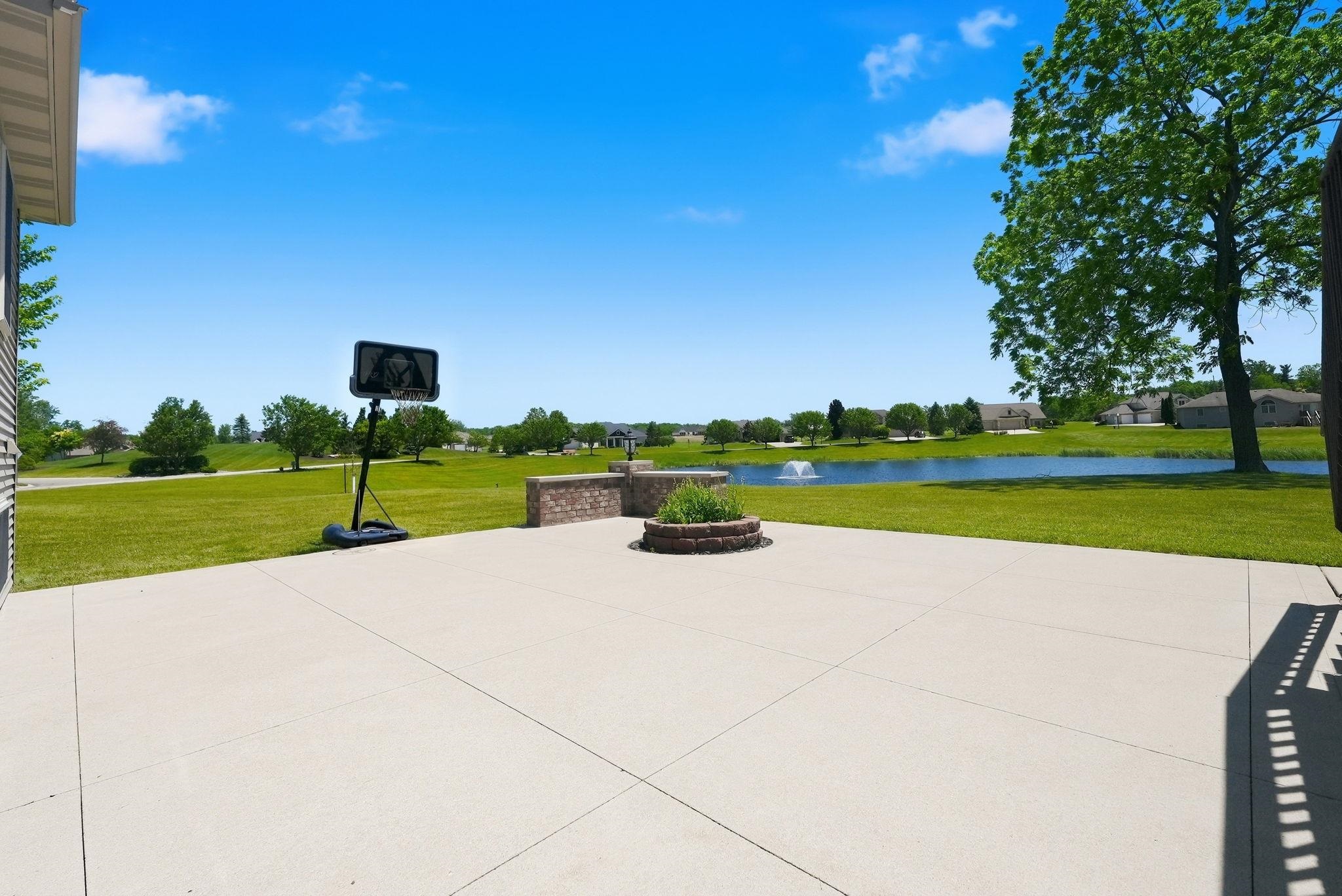 Expansive concrete patio with a stone fire pit, bordered by a low brick wall, overlooking a large grassy area and a pond with a fountain