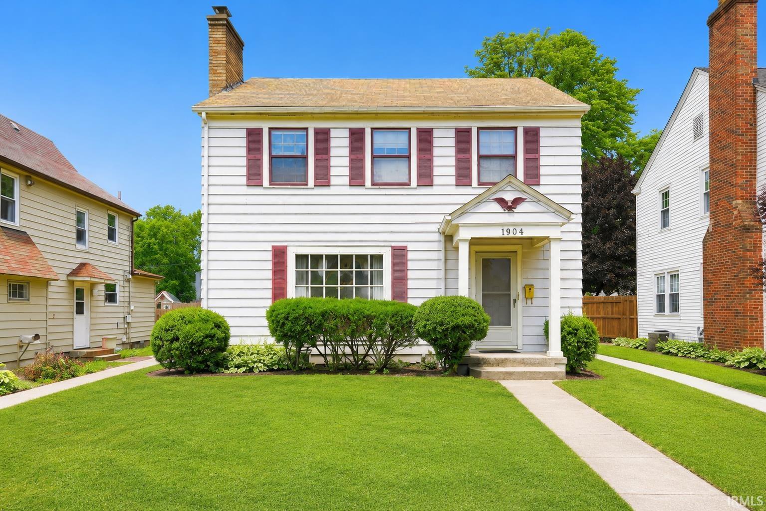 Two-story white siding residence featuring red window shutters, a covered entry with columns, a prominent front window with multiple panes, and a brick chimney