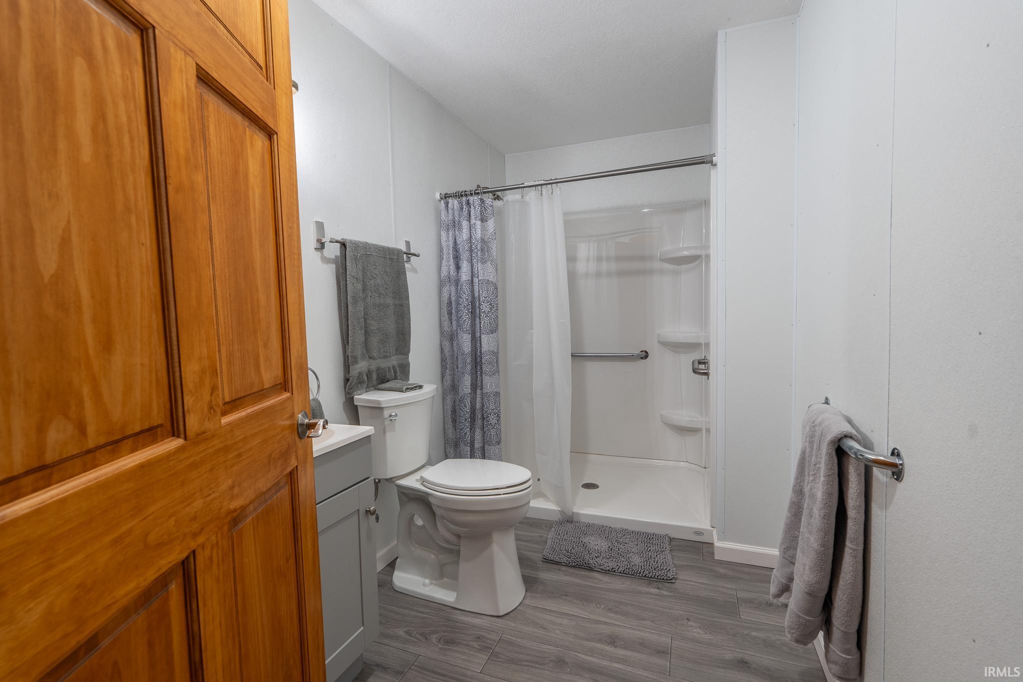 Bathroom featuring a walk-in shower with integrated shelving, wood-finish flooring, a toilet, a vanity with a white countertop, and a wood-panel door
