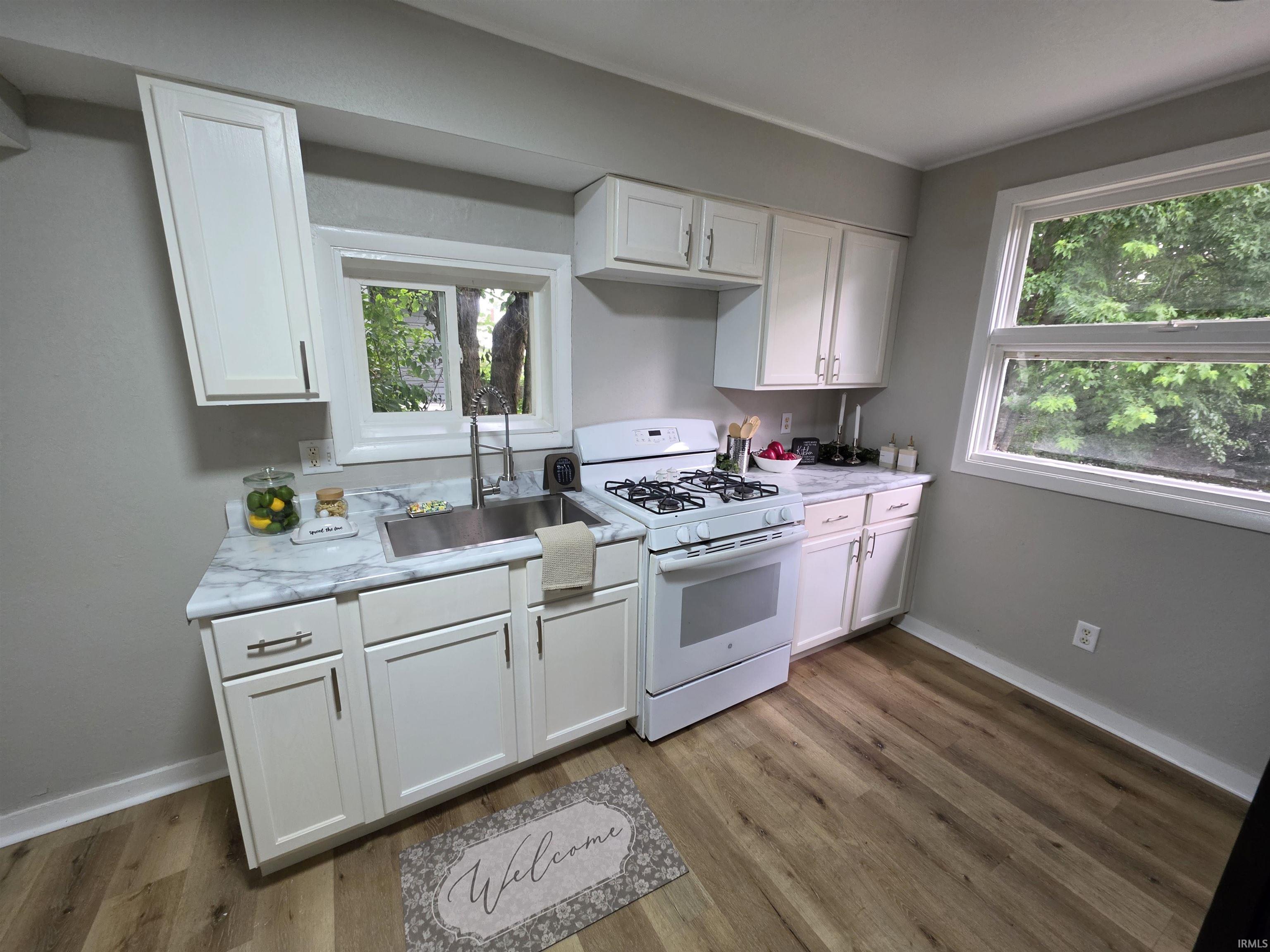 Kitchen featuring white cabinetry with dark hardware, a stainless steel sink, a white gas range, light-toned wood-finish flooring, and a large window overlooking exterior greenery