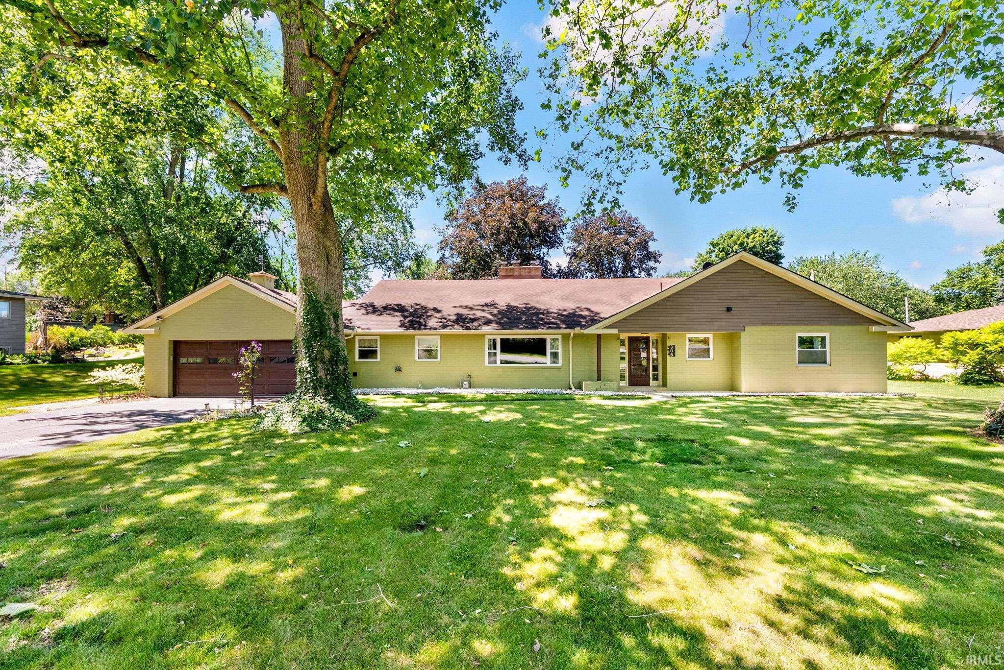 Ranch-style residence featuring a prominent front lawn, mature shade trees, an attached garage with a wood-finish door, and a covered entryway