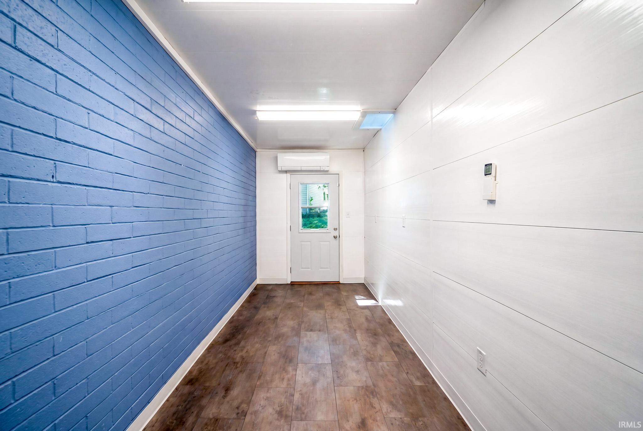 Interior space featuring a blue brick accent wall, white shiplap wall, wood-finish flooring, fluorescent lighting, and a white exterior door with glass panes