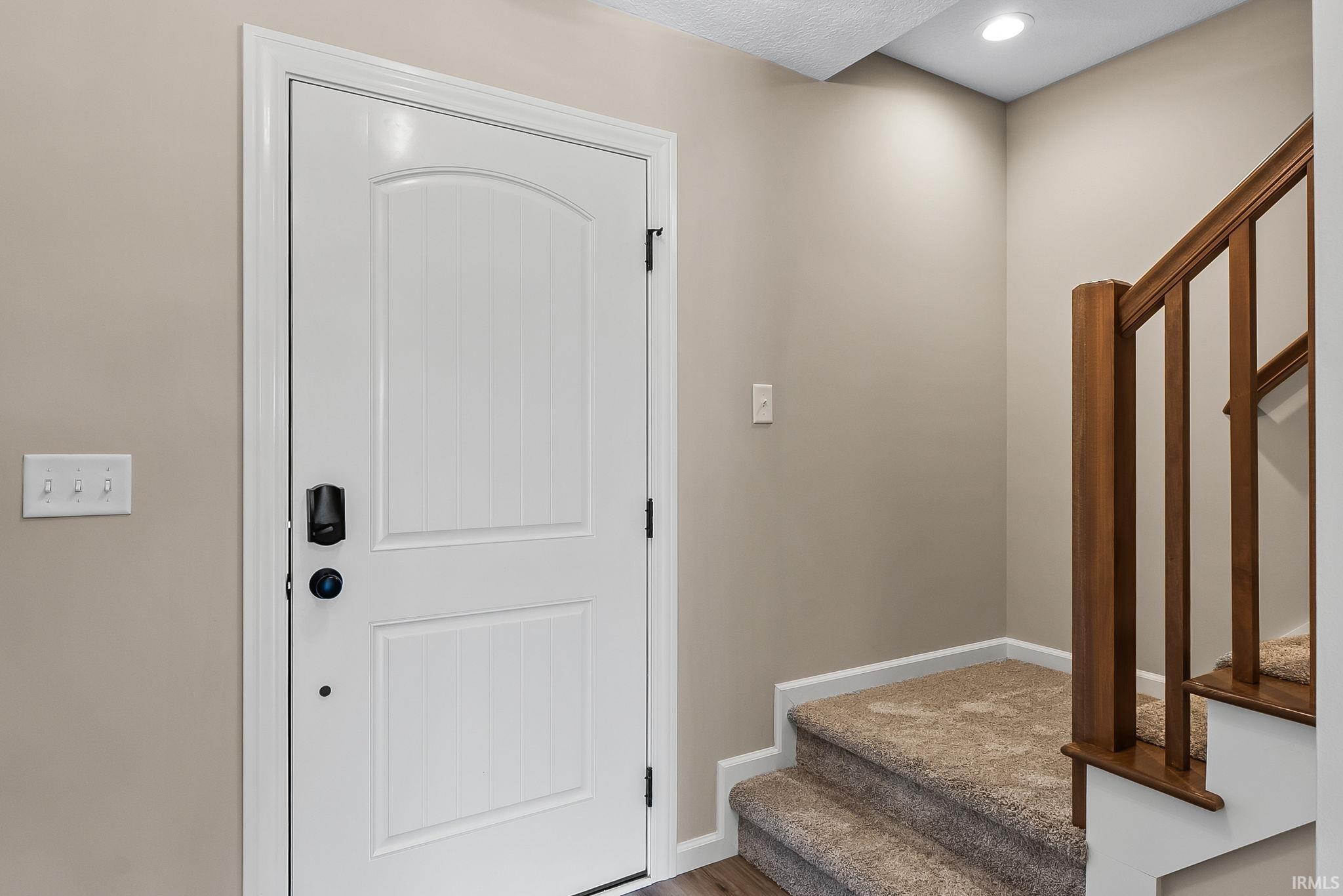 White paneled door with smart lock entry, flanked by a light switch panel, and a wood-finish staircase with carpeted treads