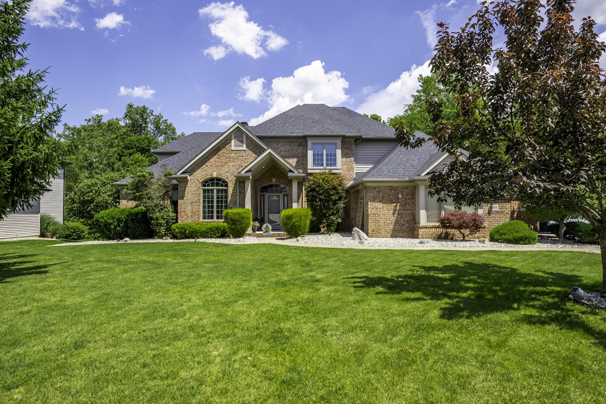 Expansive front lawn with mature trees frames a brick and siding exterior featuring a prominent arched window, symmetrical columns, and a dark shingled roof