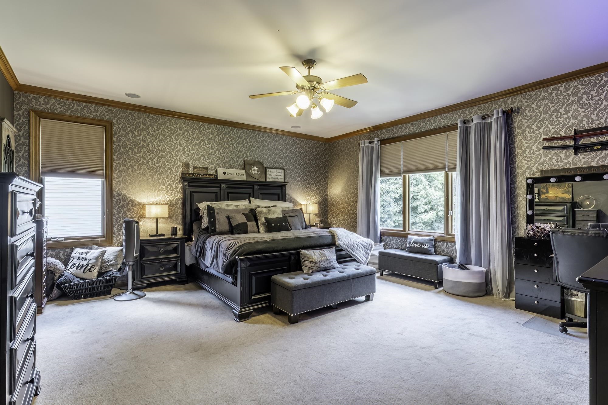 Spacious carpeted room featuring two large windows with blinds, a ceiling fan with integrated lighting, patterned wallpaper, and wood trim