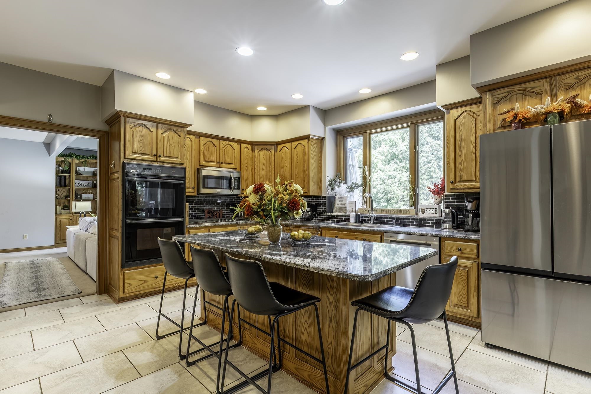 Kitchen featuring natural wood cabinetry, a granite-topped island with seating, stainless steel appliances, a dark tile backsplash, and light-toned tile flooring
