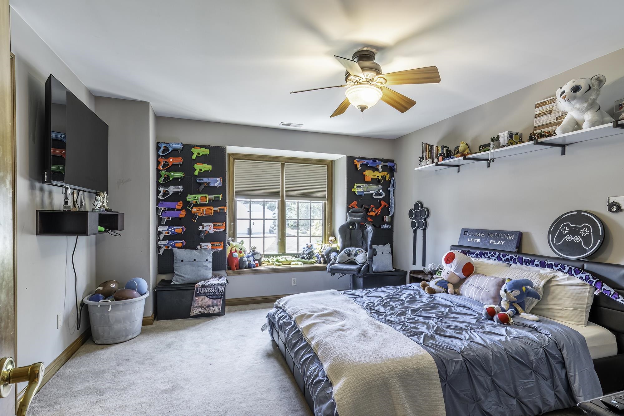Spacious bedroom featuring light-toned carpet flooring, a ceiling fan with integrated lighting, and a large window with blinds