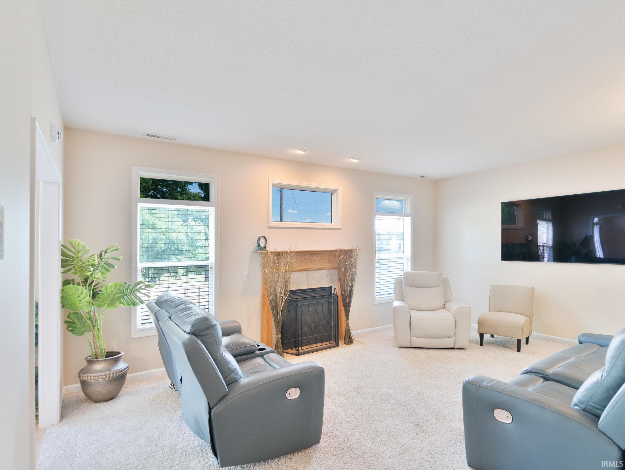 Light-colored carpeted living room featuring a fireplace with a wood mantel, multiple windows with blinds, and recessed lighting
