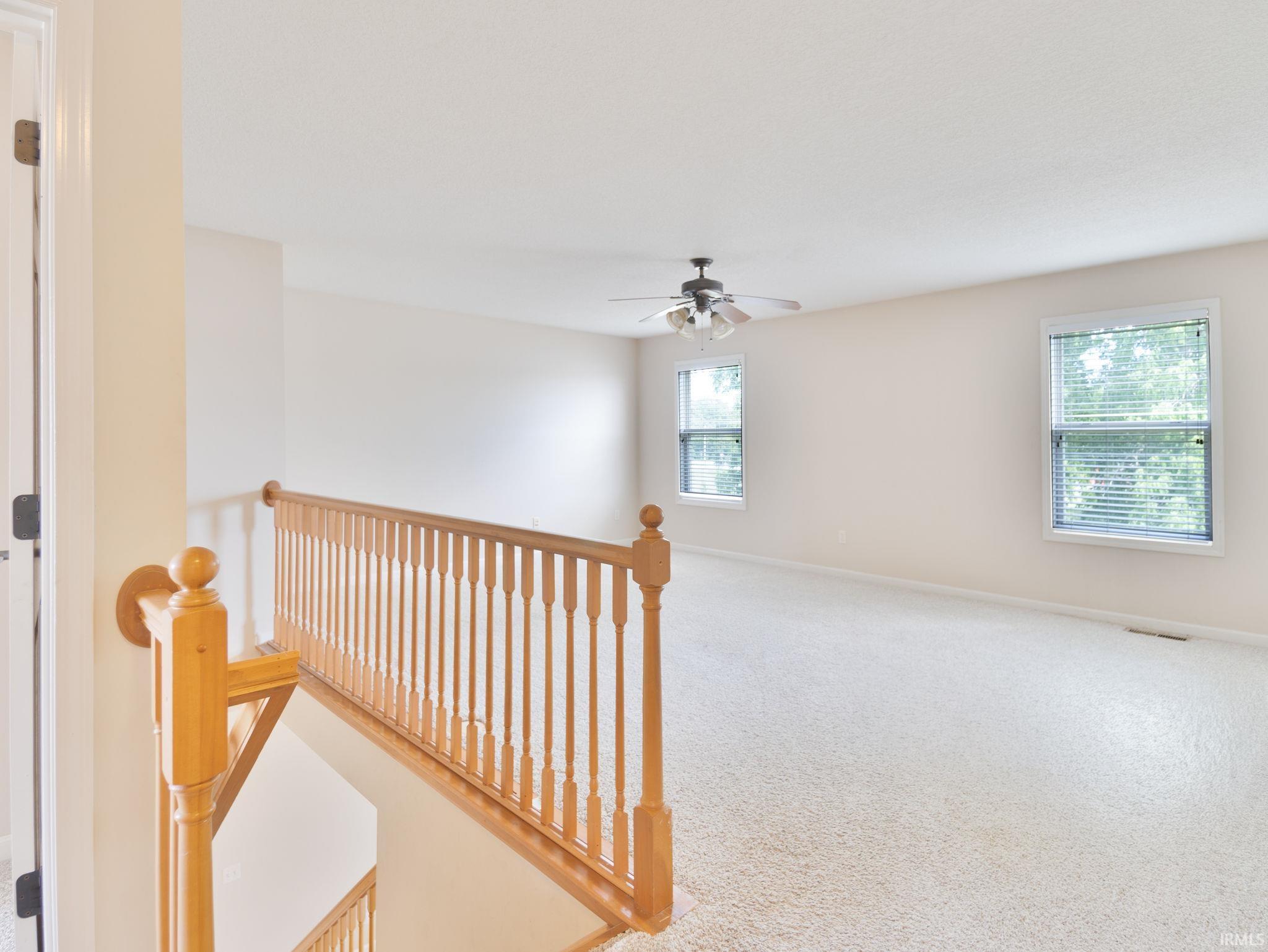 Carpeted loft area featuring a natural wood-finish railing, two windows with horizontal blinds, and a ceiling fan