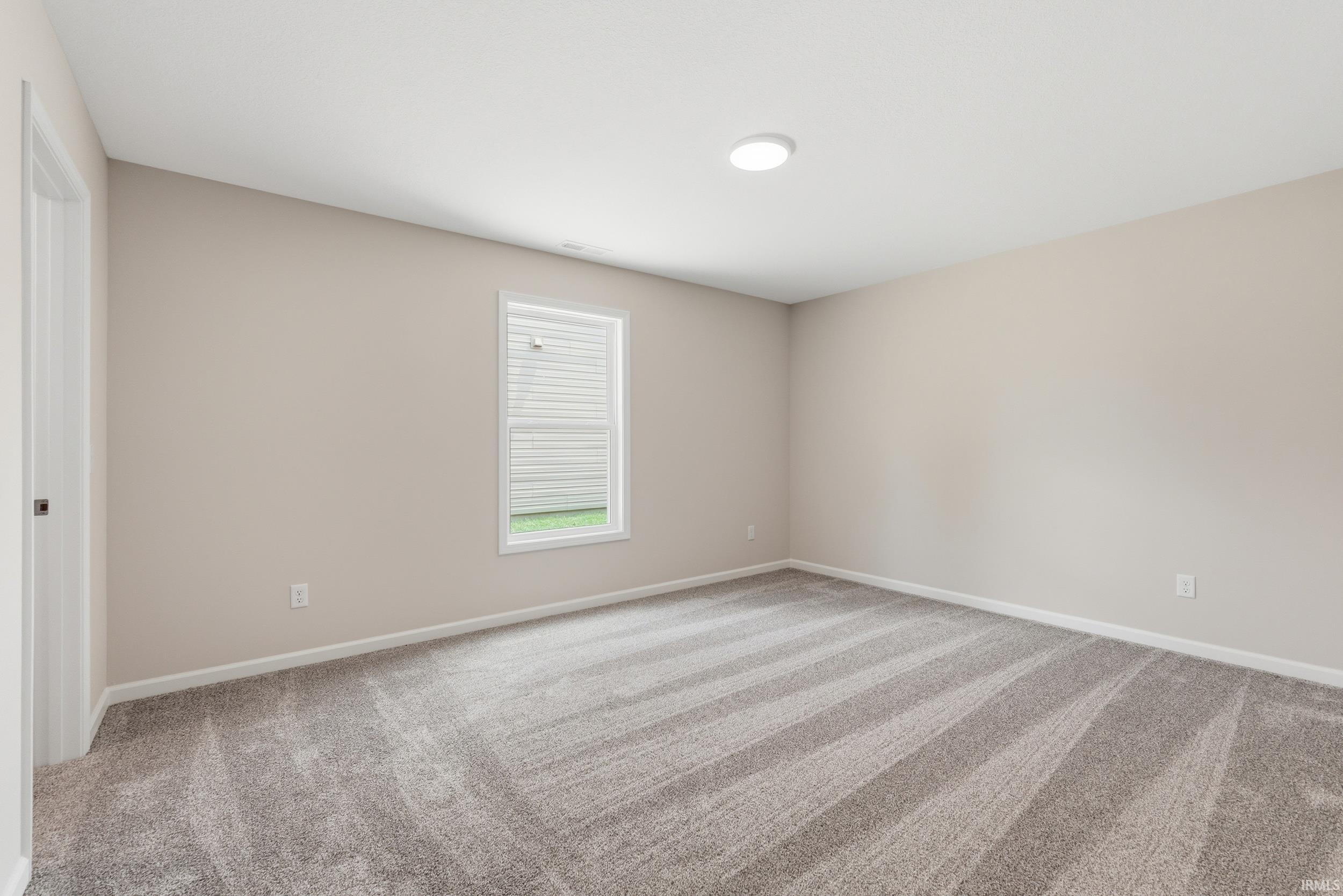 Neutral-toned wall paint with white trim, gray patterned carpet, a single window with blinds, and a flush-mount ceiling light fixture