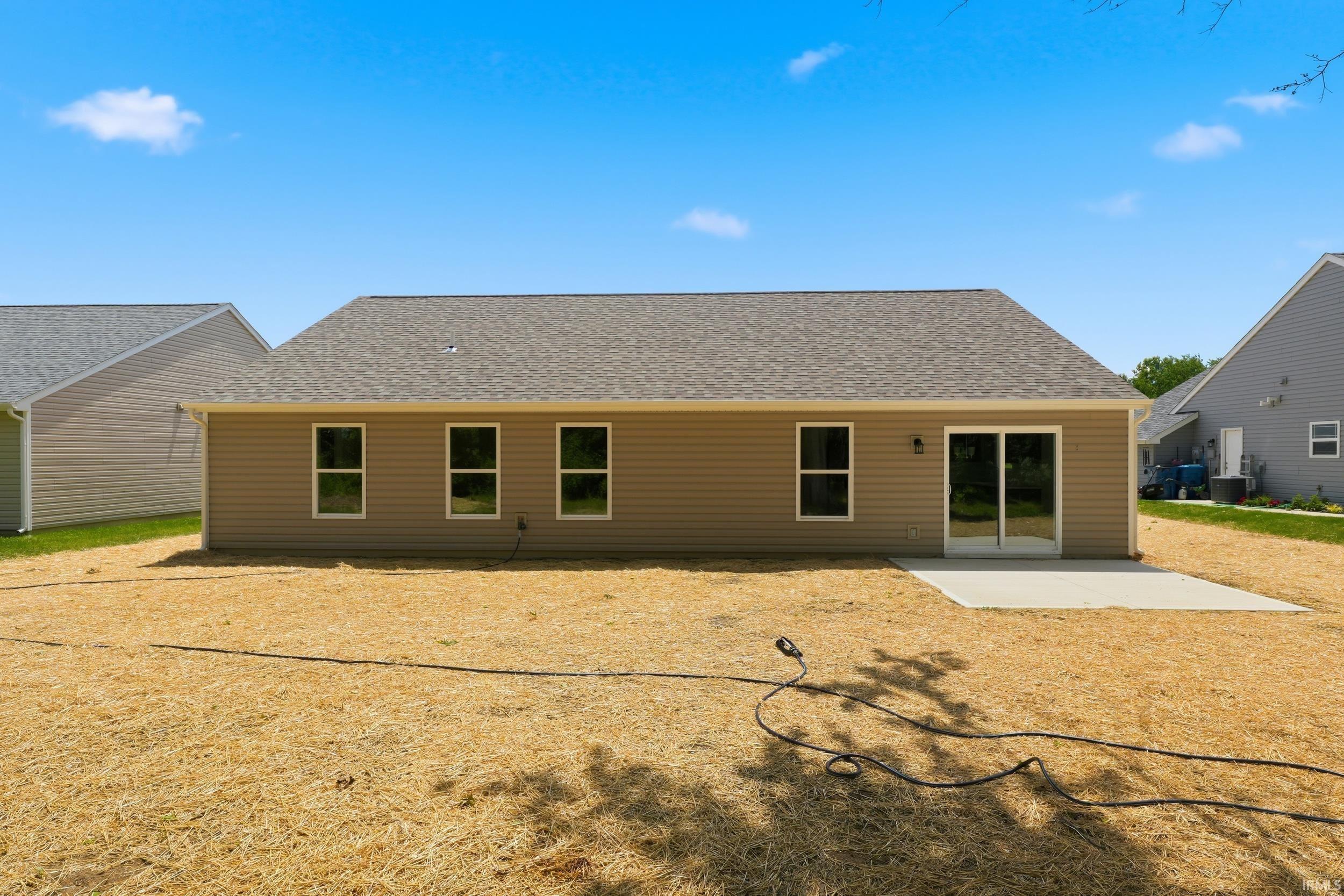Rear exterior featuring tan horizontal siding, a gray shingle roof, multiple windows, and a concrete patio