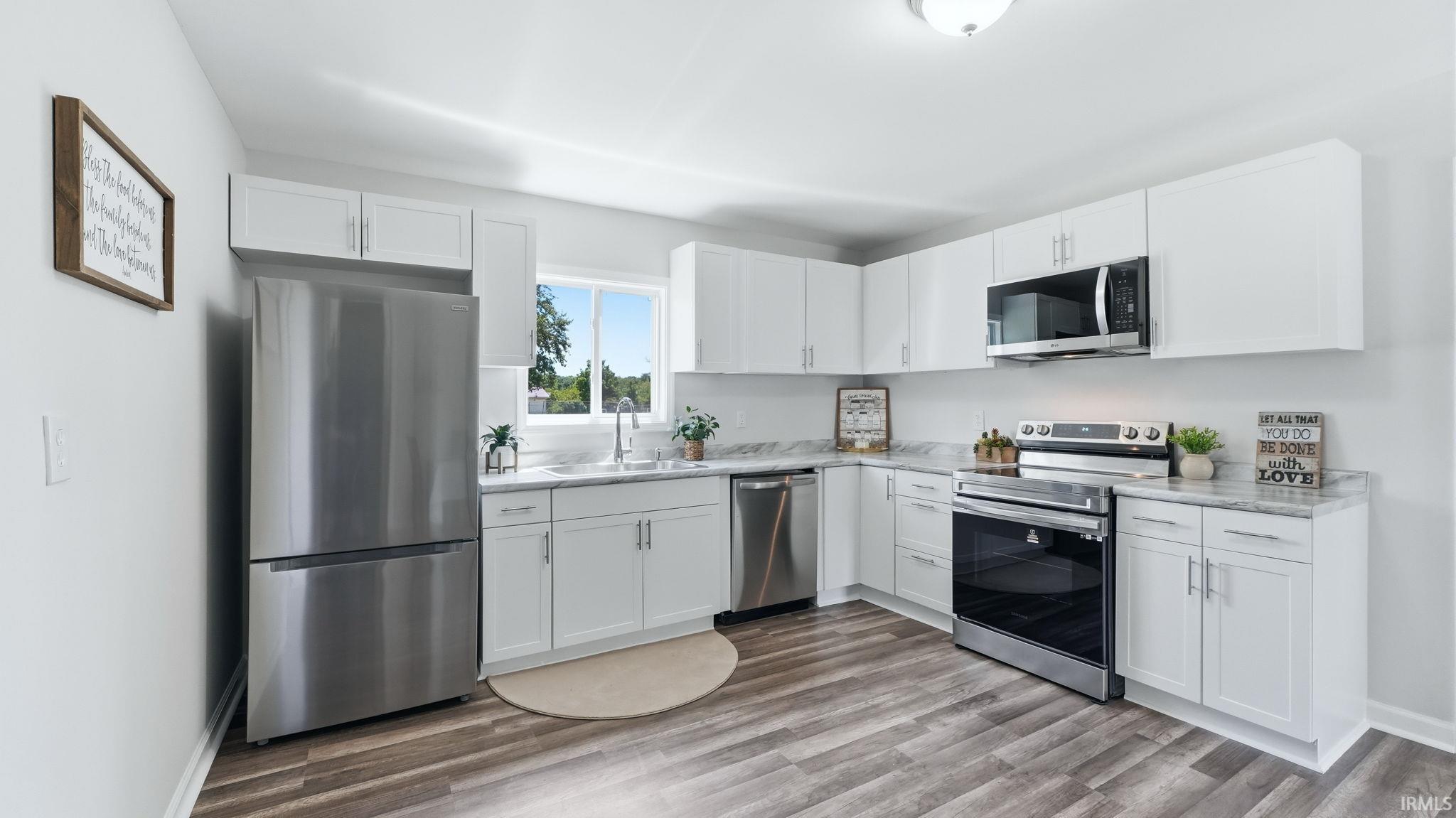 Modern kitchen featuring white cabinetry, stainless steel appliances, light-toned countertops, wood-finish flooring, and a large window above the sink