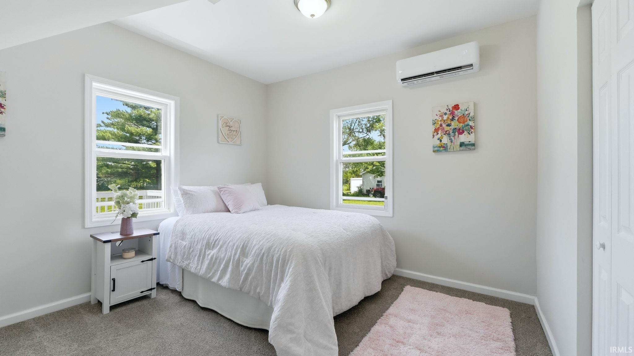 Bedroom featuring neutral gray wall paint, gray carpet flooring, dual windows with white trim, and a wall-mounted mini-split HVAC unit