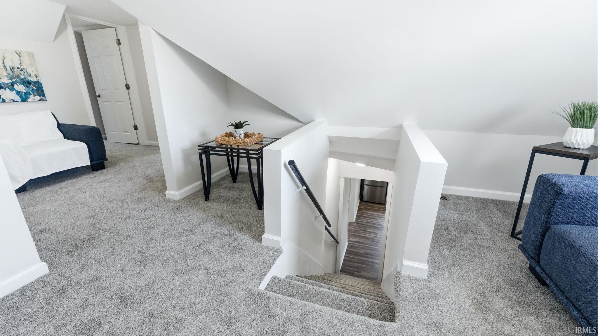 Carpeted upper-level space featuring a white door, sloped ceilings, and access to a lower level via carpeted stairs with a black handrail