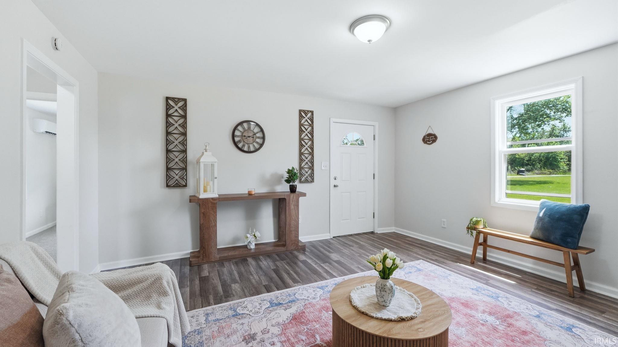 Spacious living area featuring wood-finish flooring, ample natural light from a prominent window, a white entry door with decorative glass, light-colored walls, and a ceiling-mounted light fixture