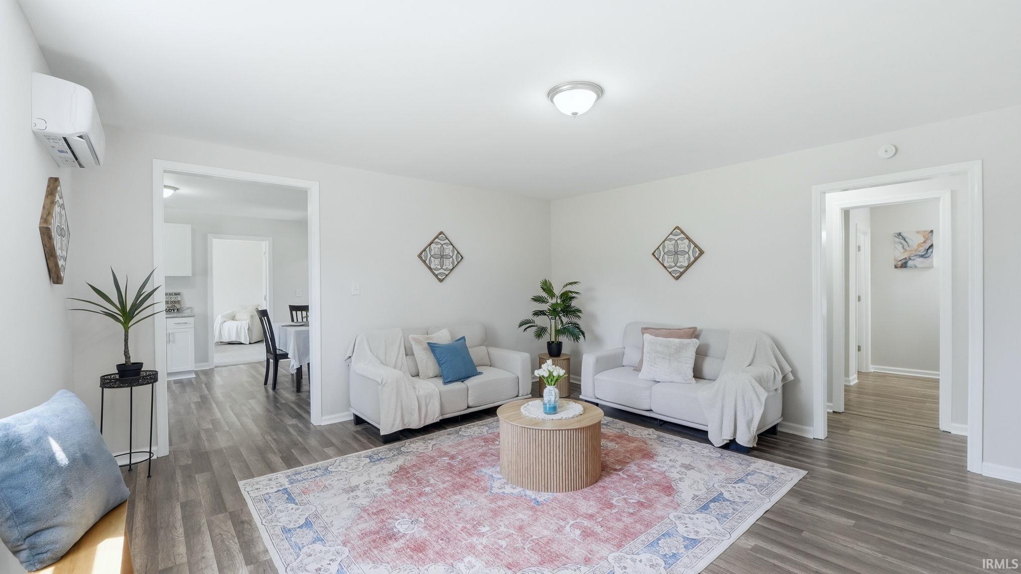 Spacious living area featuring wood-finish flooring, light neutral wall tones, a ceiling-mounted light fixture, and an in-wall air conditioning unit