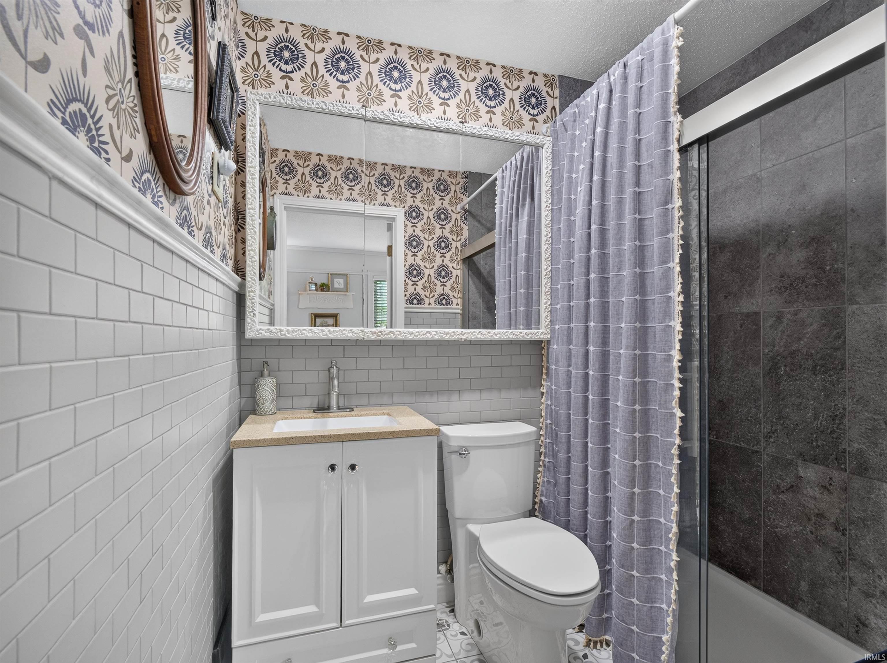 Bathroom featuring a white vanity with a light-toned countertop, a frameless shower with dark-toned wall tiling, white subway tile wainscoting, and decorative wallpaper