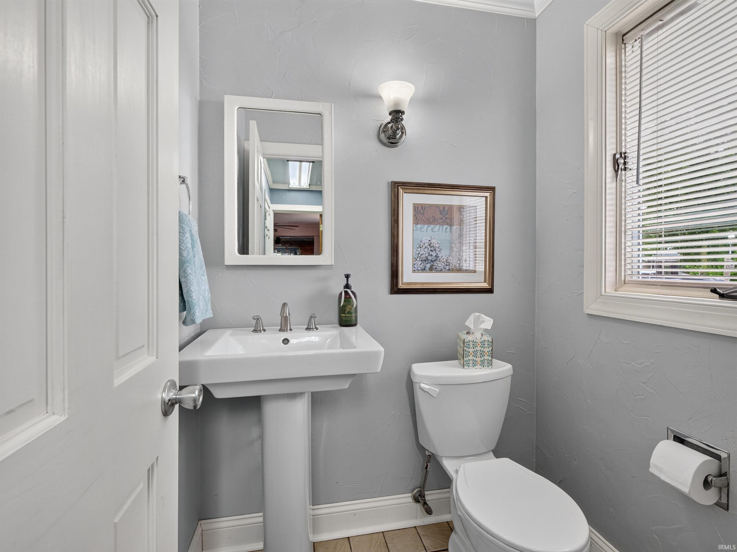 Pedestal sink with polished chrome fixtures, framed mirror, wall-mounted sconce, and recessed window