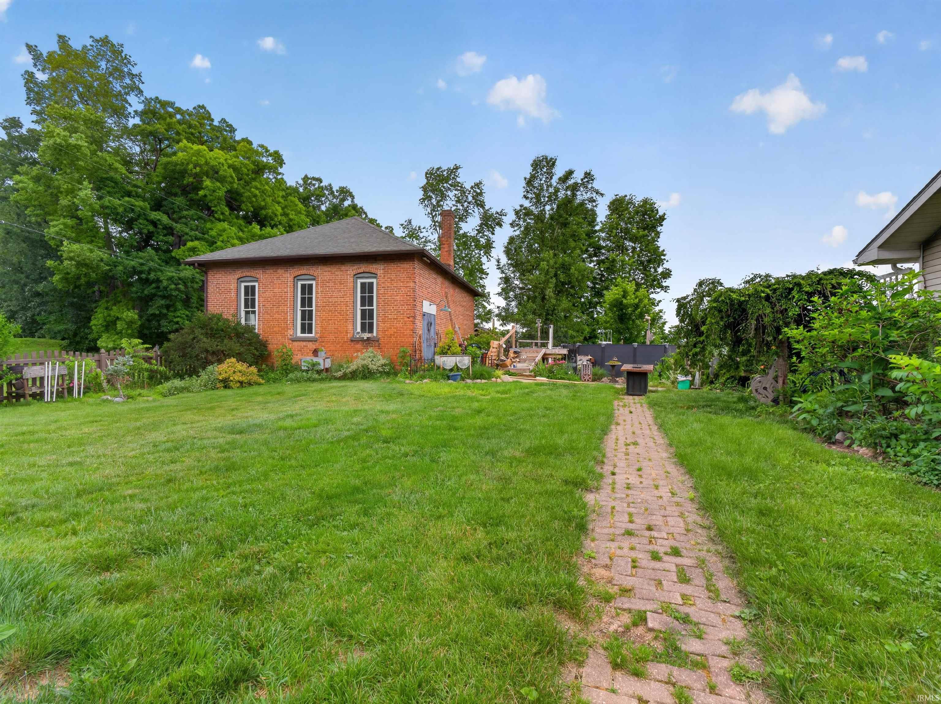 Expansive green lawn with a brick paver pathway leading to a red brick building featuring multiple windows, a prominent chimney, and mature perimeter trees