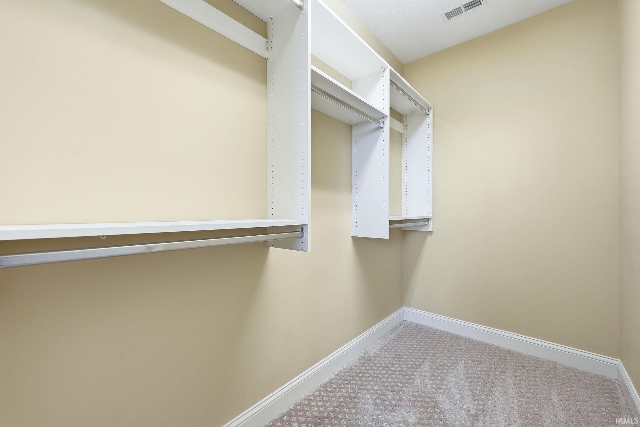 Walk-in closet featuring built-in white shelving, hanging rods, light beige wall paint, and patterned carpeting with white baseboards